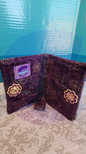 A purple wallet with gold lotus patten standing up with a gold vintage flower push bottom in the center. The wallet is open in a book position.