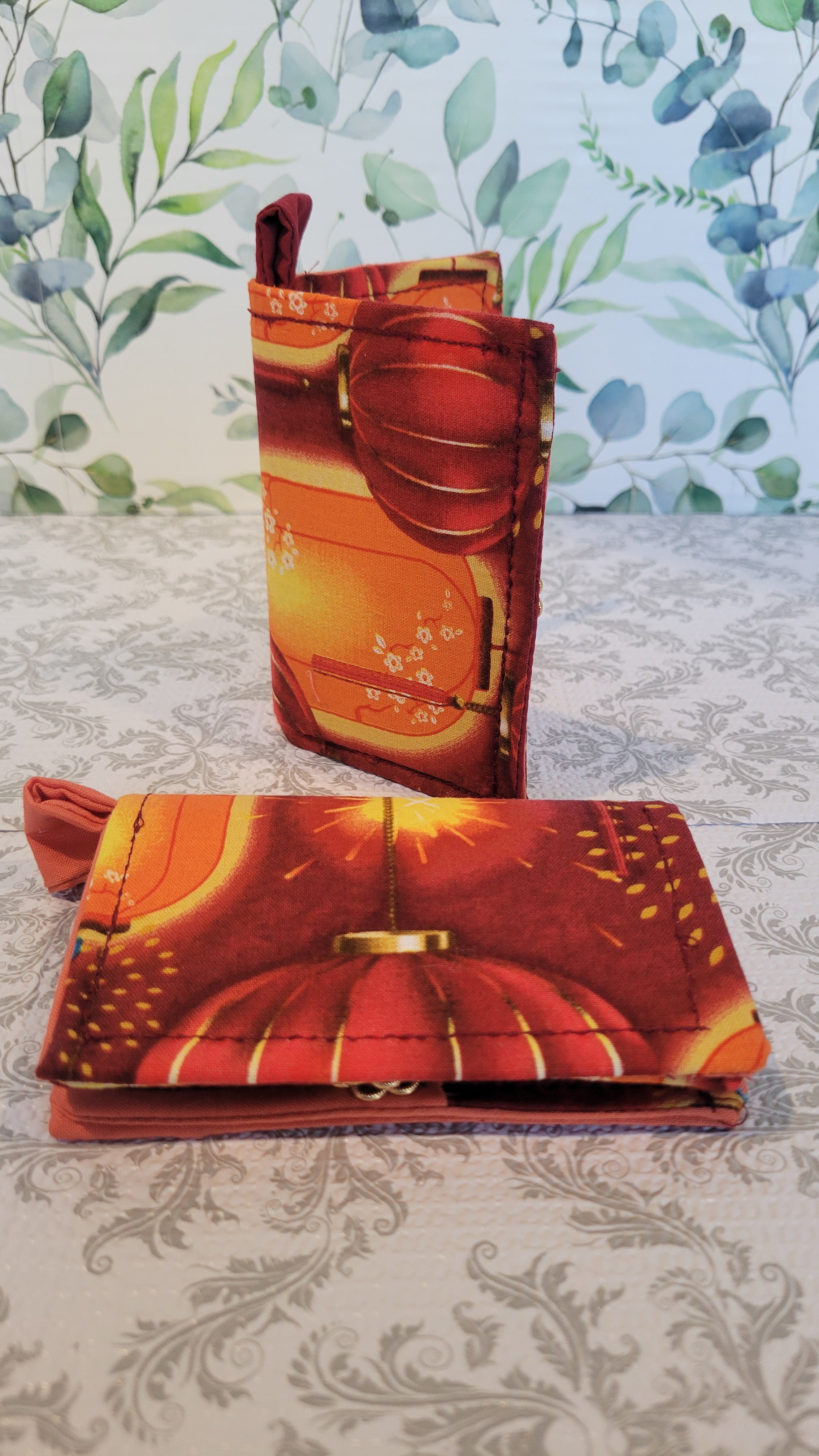 Two Orange Lantern Design Wallet on red fabric. Standing on top of a crazy lace design and a leave pattern background. One is face front and the second is standing like a book open on the middle.