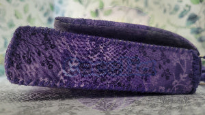 a side picture of a purple floral patten clutch purse on a textured surface. Attach is two sliver hook and adjustable crochet strap in purple.