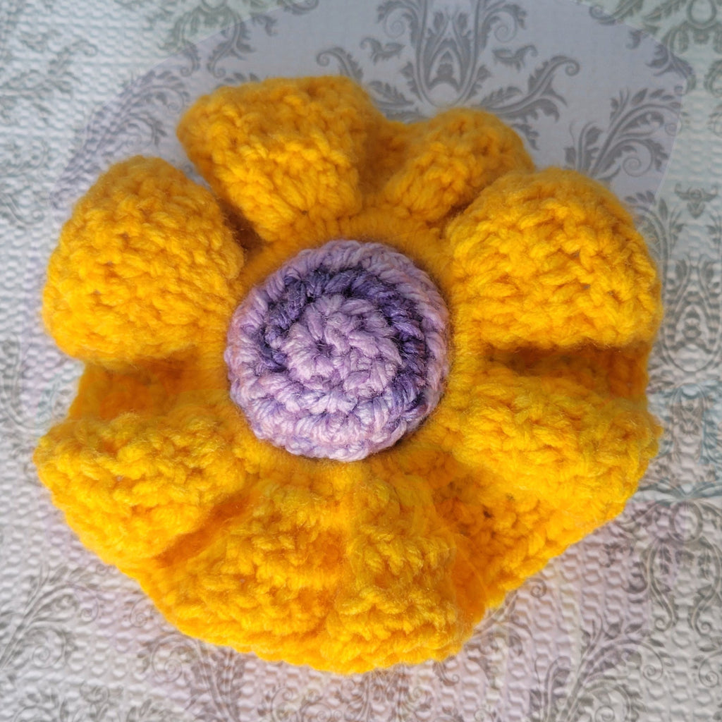 Crocheted yellow scrunchie with a purple ball center on a blue and patterned surface