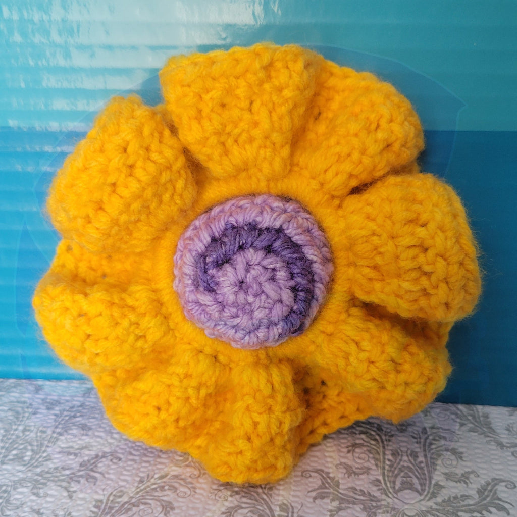 Crocheted yellow scrunchie with a purple ball center on a blue and patterned surface