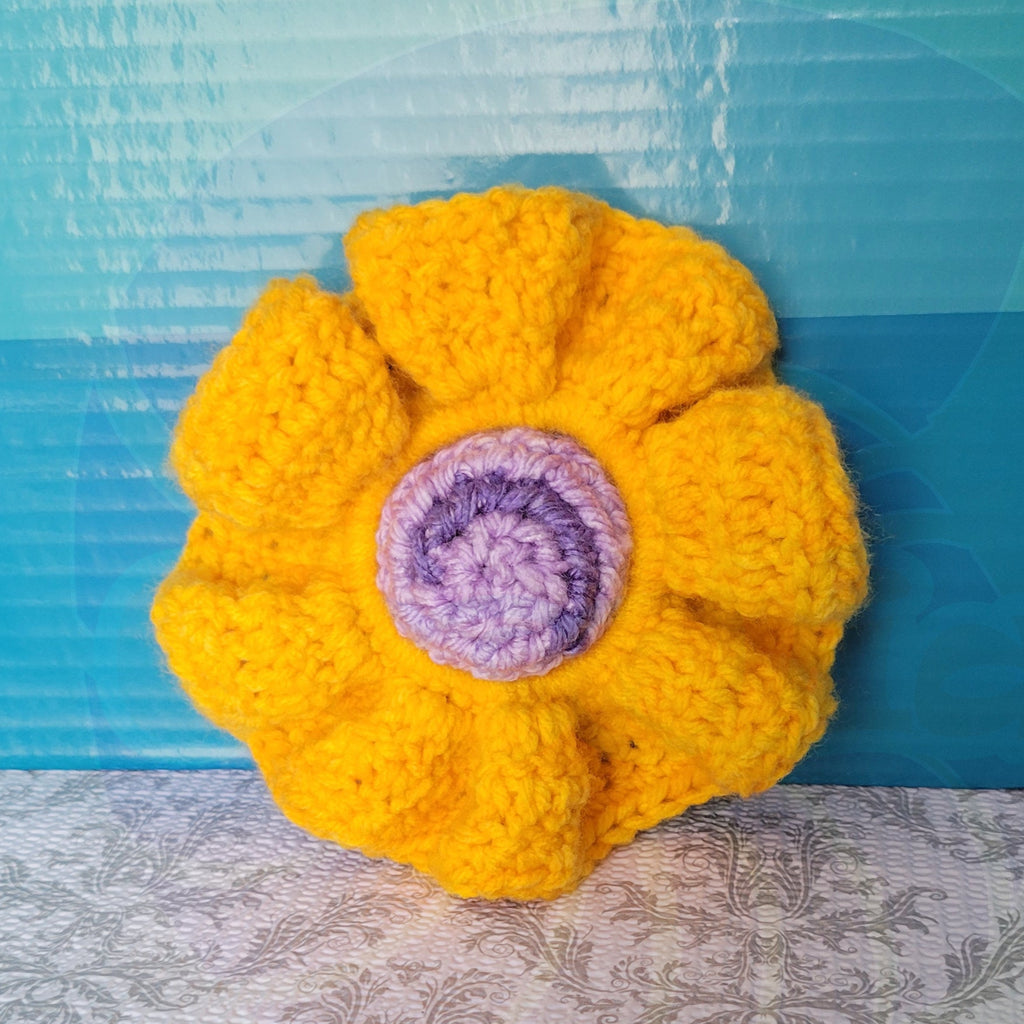 Crocheted yellow scrunchie with a purple ball center on a blue and patterned surface