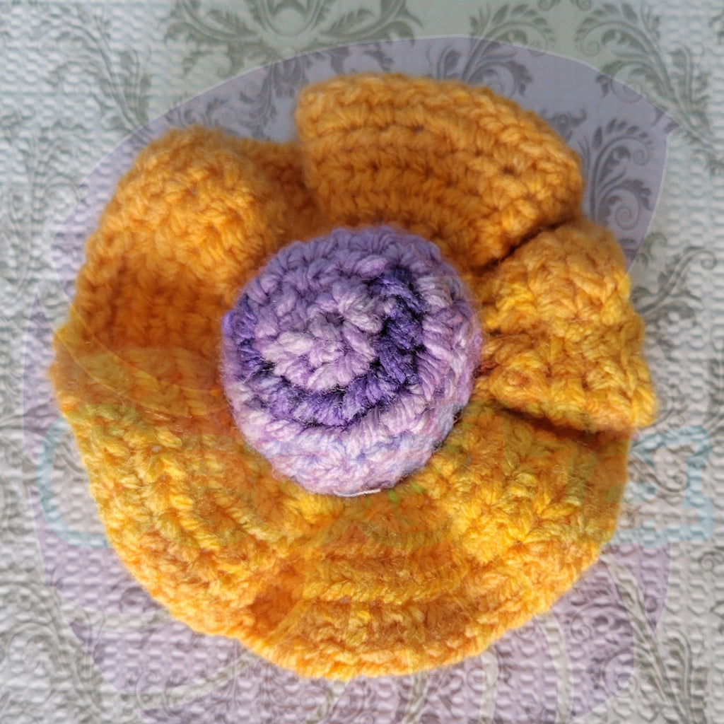 Crocheted yellow scrunchie with a purple ball center on a blue and patterned surface