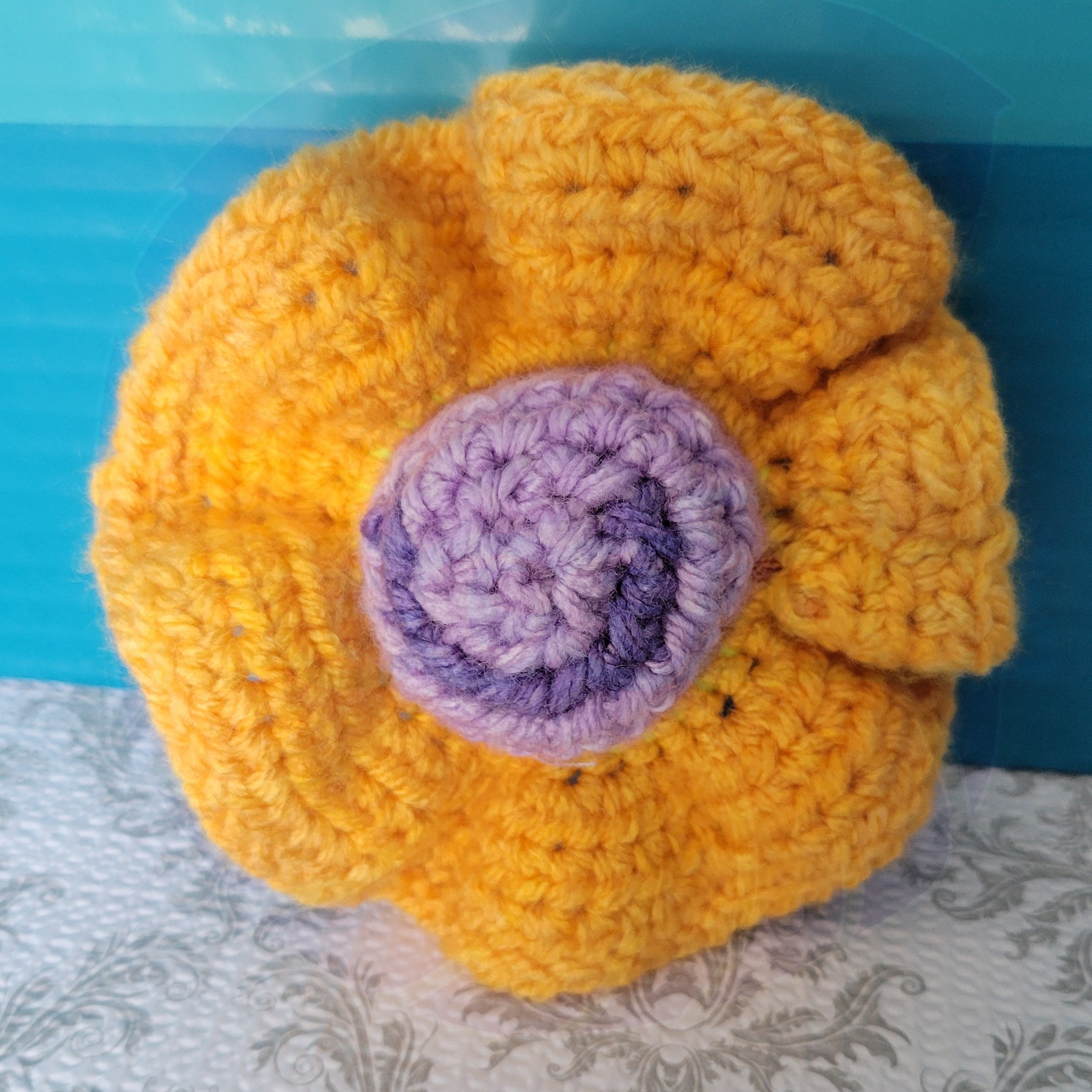 Crocheted yellow scrunchie with a purple ball center on a blue and patterned surface