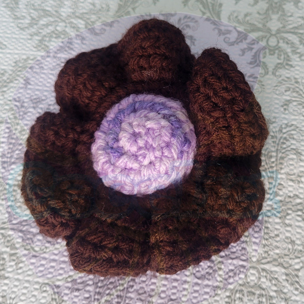 Brown crocheted scrunchie with a purple center ball on a textured surface.