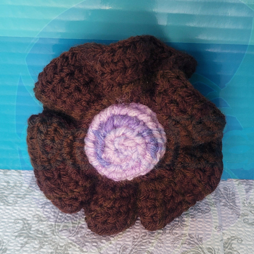 Brown crocheted scrunchie with a purple center ball on a textured surface.