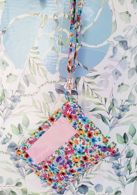 colorful Floral-patterned pouch purse in white with a light pink pocket on a textured white background. Attach to a pink metal hook is the hand size strap which is hanging on its hook.