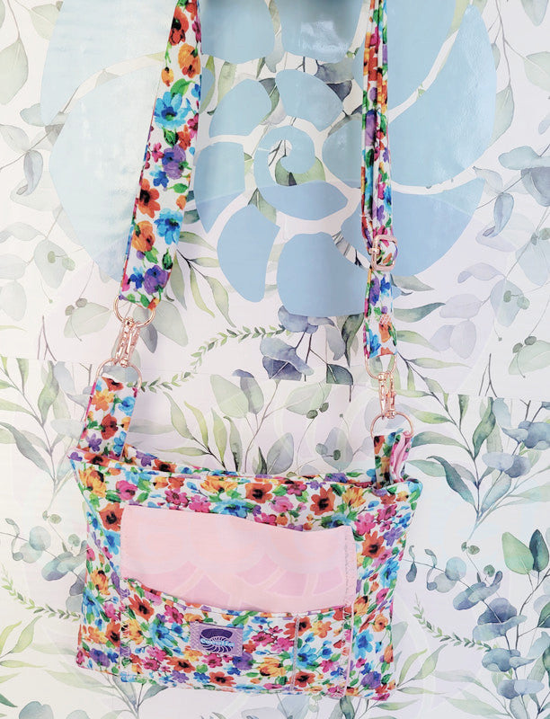 colorful Floral-patterned pouch purse in white with a light pink pocket on a textured white background. Attach to a set of pink metal hooks is the adjust strap in the same floral pattern and is handing on a hook.