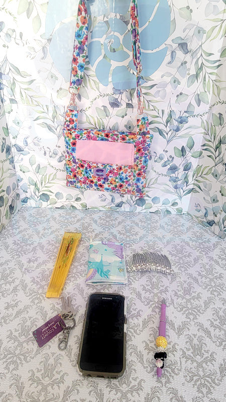 colorful Floral-patterned pouch purse in white with a light pink pocket on a textured white background. Attach to a pink metal hook is the adjustable size strap in the same floral pattern. In Front of the hanging purse is a yellow fan, a unicorn, a pair of white hair combs, a group of keys, a cell phone and a pen. This photos show how much fits inside the purse.