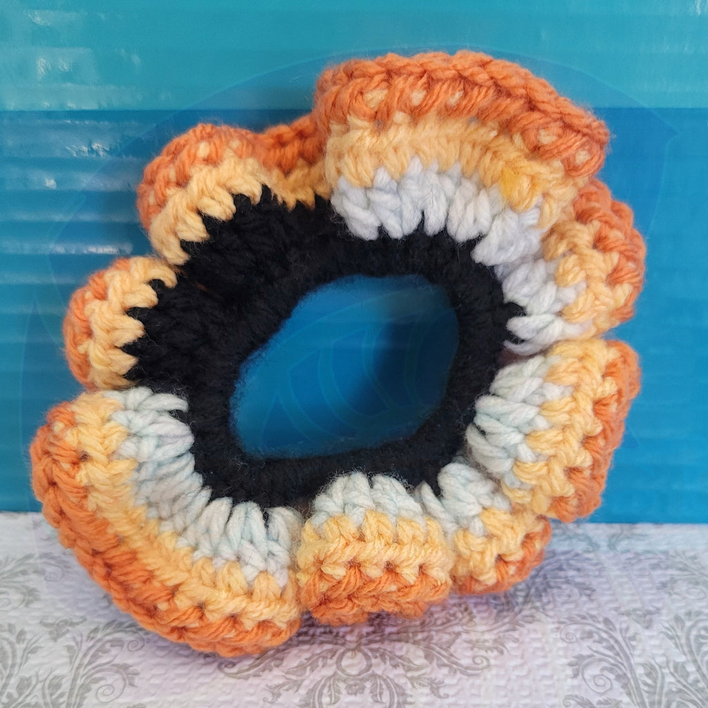 Crocheted scrunchie with orange, black, and white colors on a textured surface with a blue background.