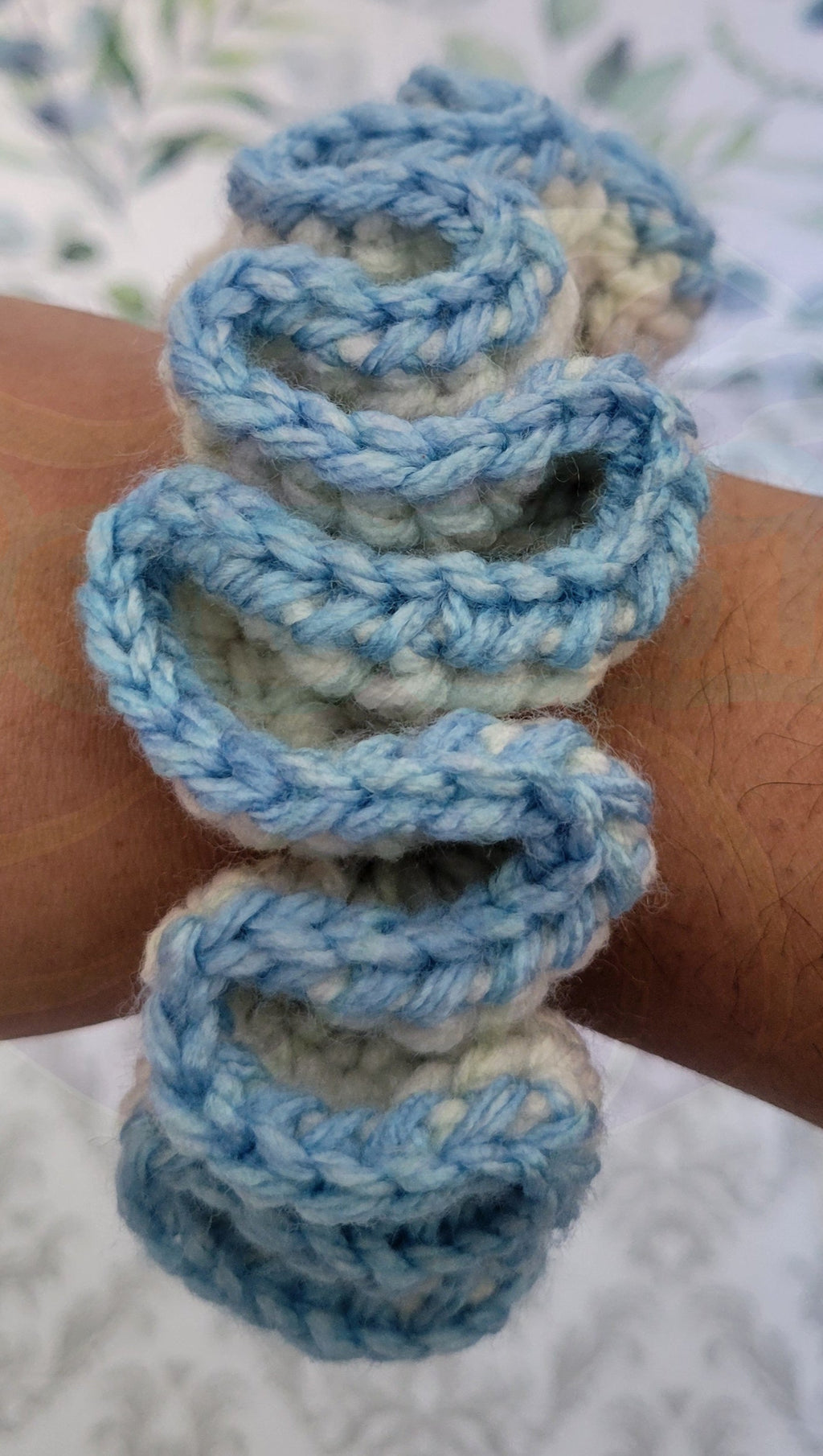 Blue and beige crochet scrunchie on a person's wrist with a floral patterned background