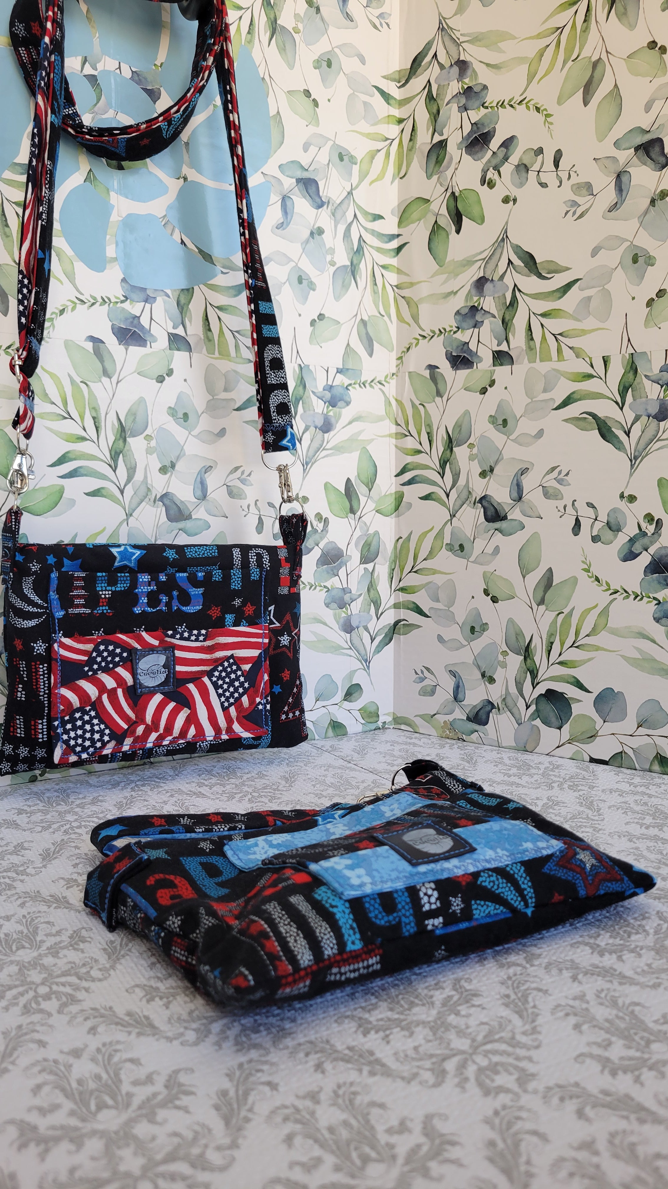Two matching purses set featuring a small shoulder bag with a red, white, and blue American flag pattern and strap, and a folded matching pouch or case with a blue and red abstract print, displayed against a floral wallpaper background.