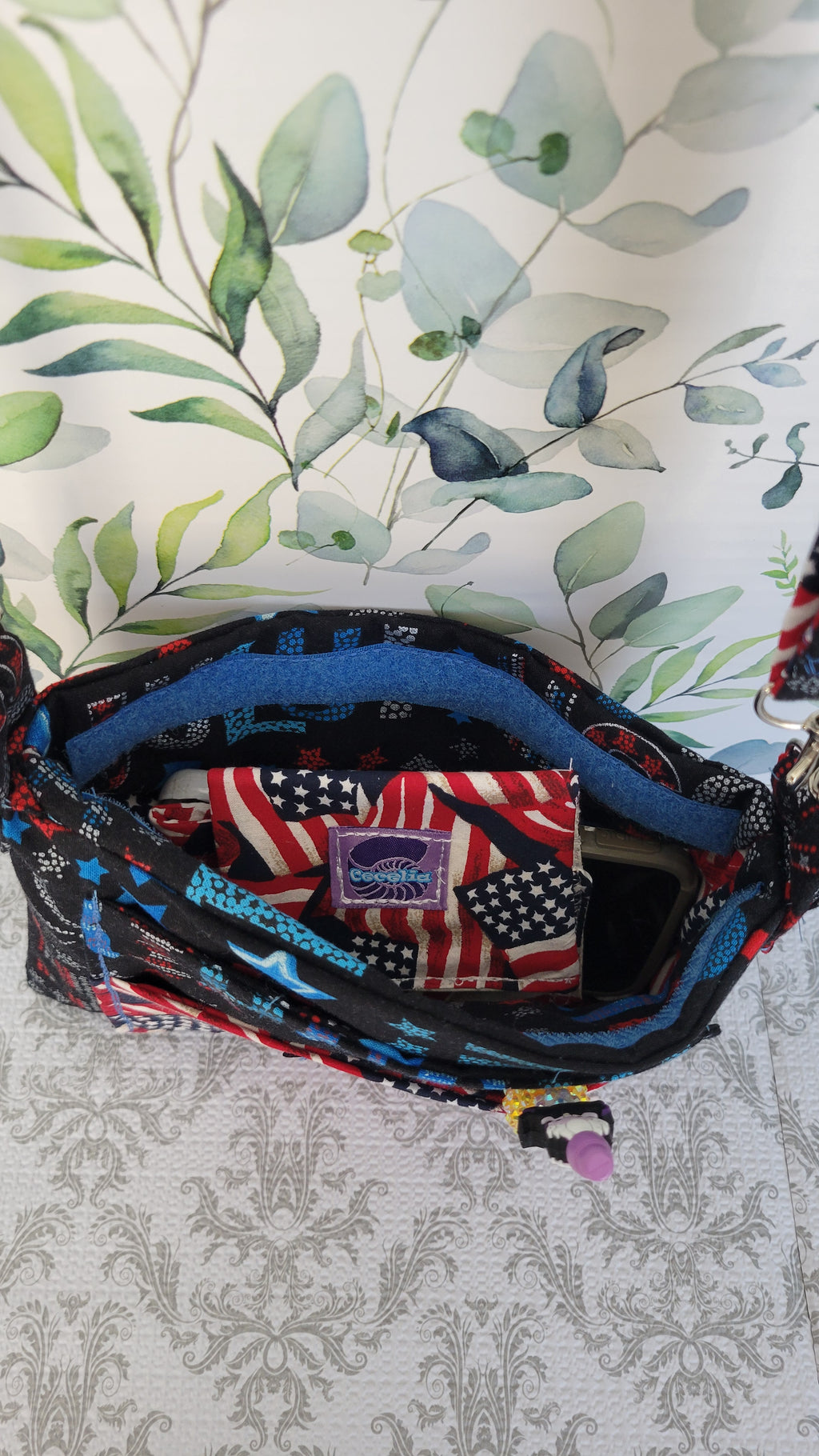 An open black patterned purse with blue lining and a red, white, and blue American flag patterned interior, photographed on a light patterned surface against a green leaf print background.