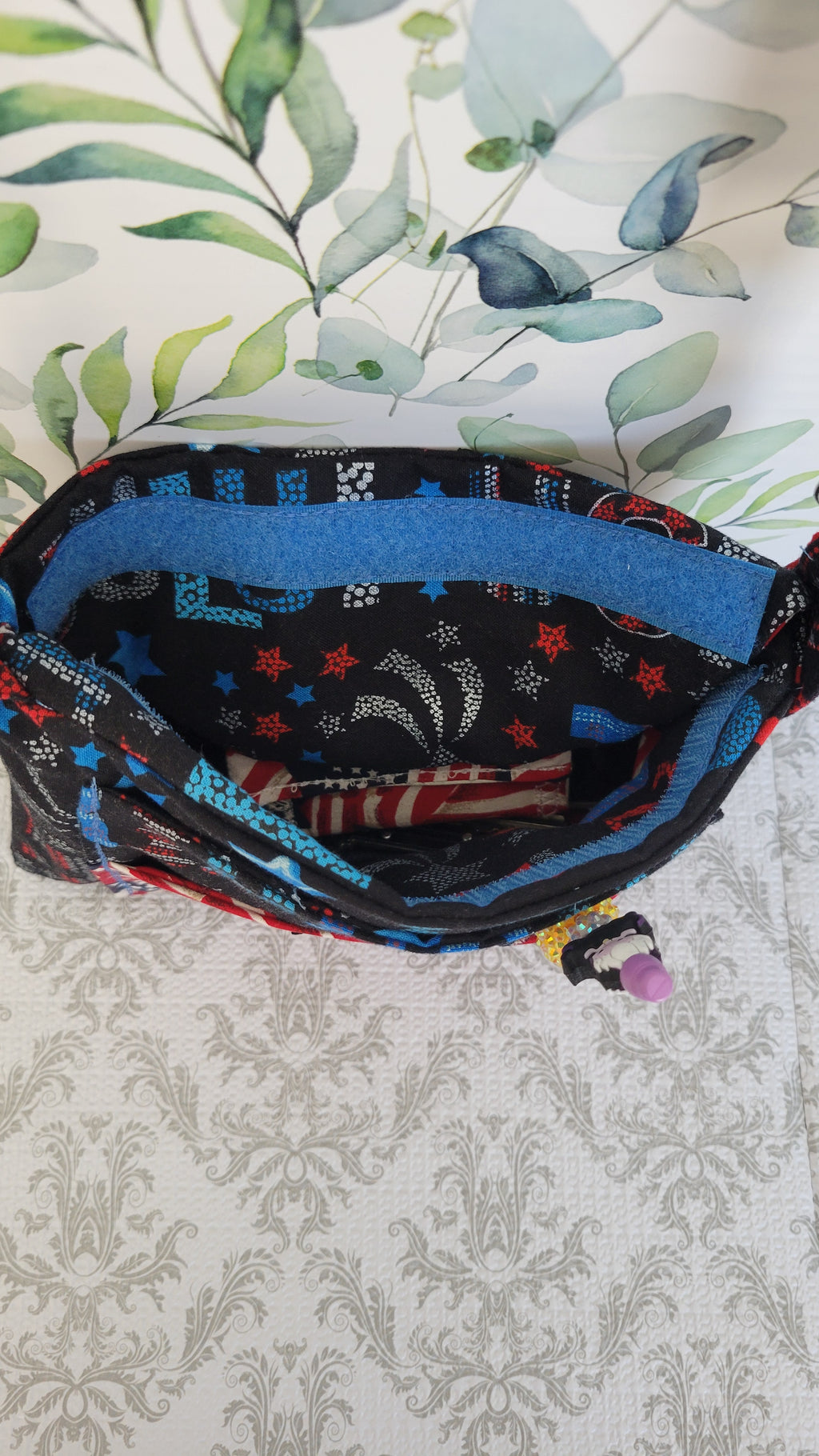 A purse with a multicolor print of stars, stripes, and geometric shapes, shown open to display its interior lining. The pouch is placed on a light-colored surface with a gray damask pattern, with a watercolor eucalyptus leaf print background visible behind it.