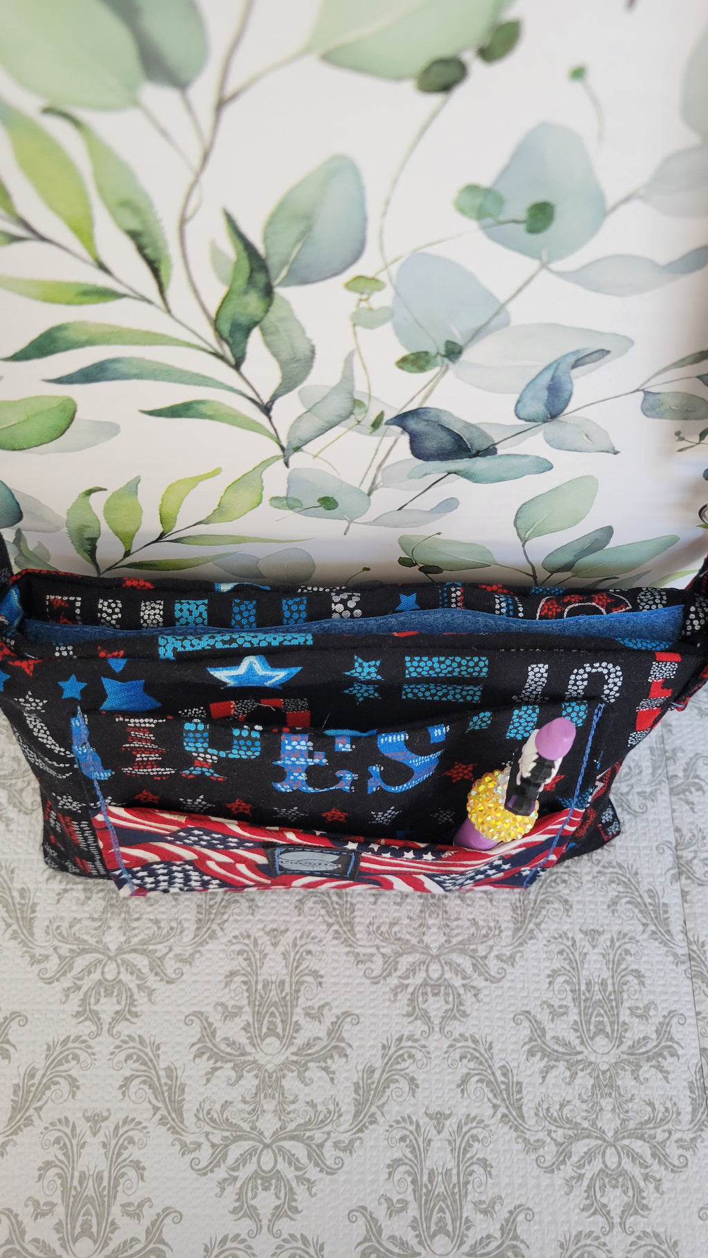 A patterned fabric bag with a black base and colorful patriotic-themed graphics, resting on a patterned surface against a leafy wallpaper background. On the front pocket is a ursula pen. This pen is not included.