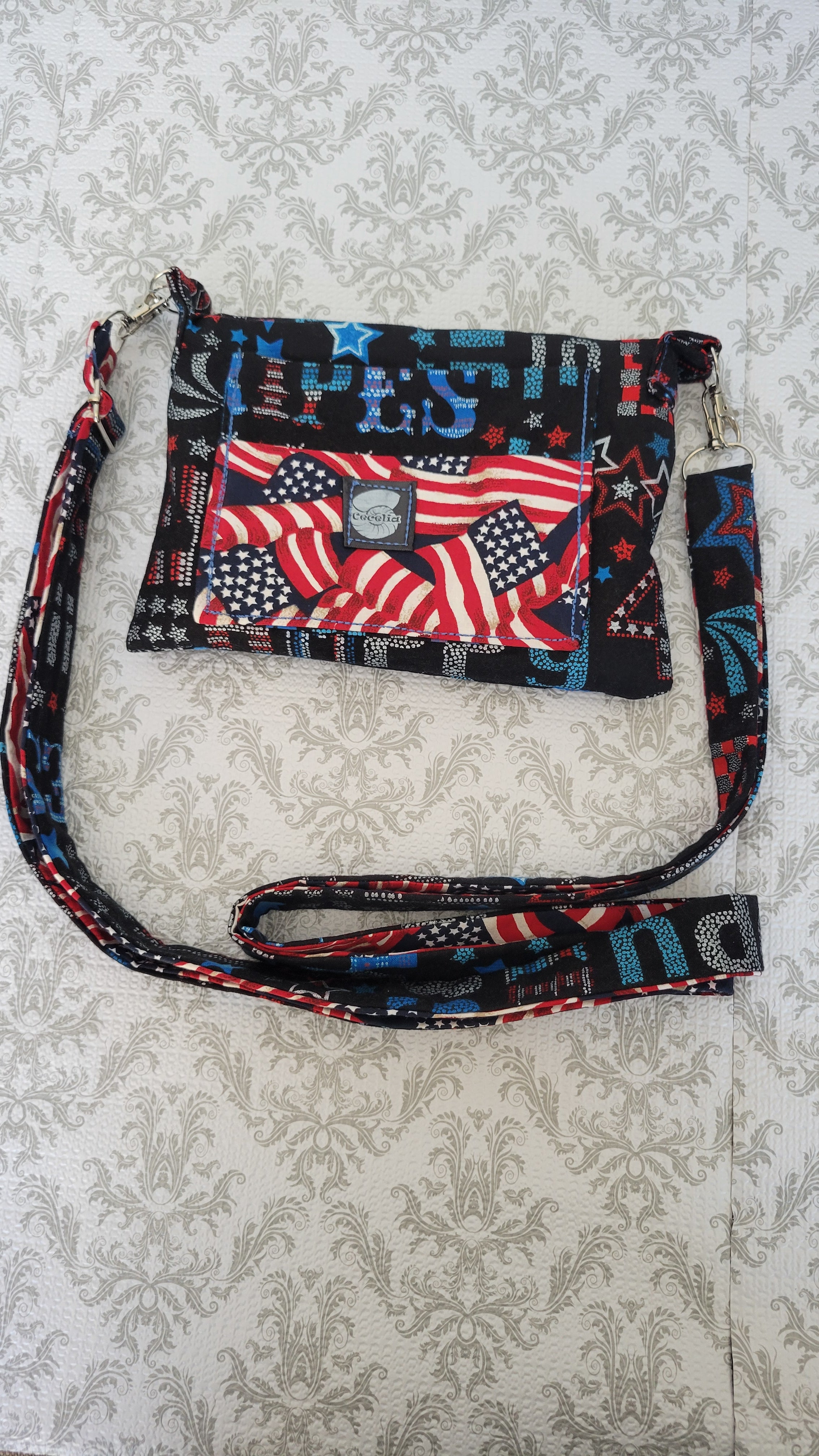 A patriotic-themed clutch purse with a matching wrist strap, featuring a red, white, and blue American flag pattern with stars and stripes, laid flat on a light beige damask-patterned background.