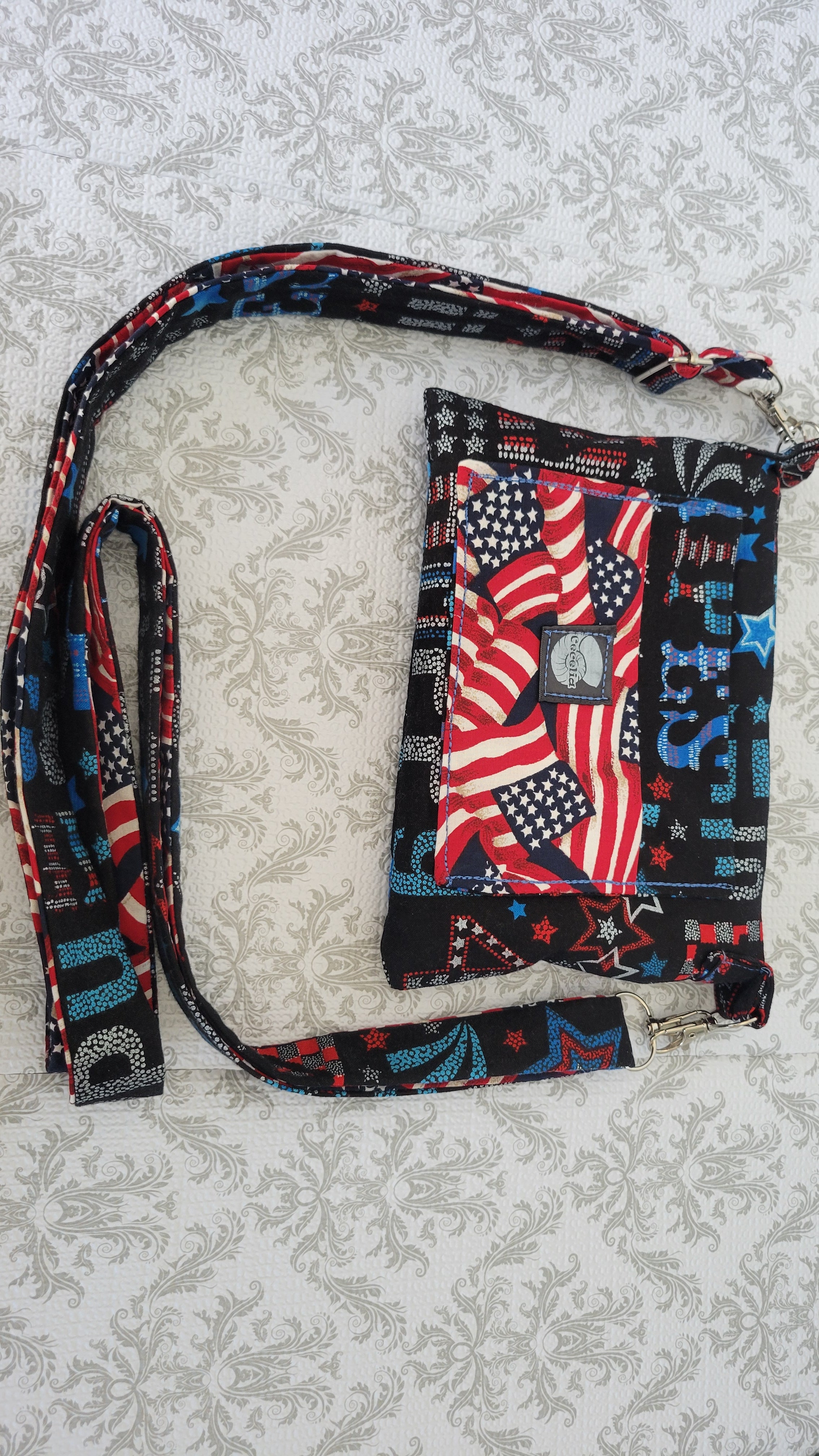 A small fabric crossbody purse with a patriotic American flag print, featuring red and white stripes, white stars on blue, and a central baseball patch. It has a matching adjustable shoulder strap and silver-tone metal hardware, laid flat on a light beige patterned fabric background.