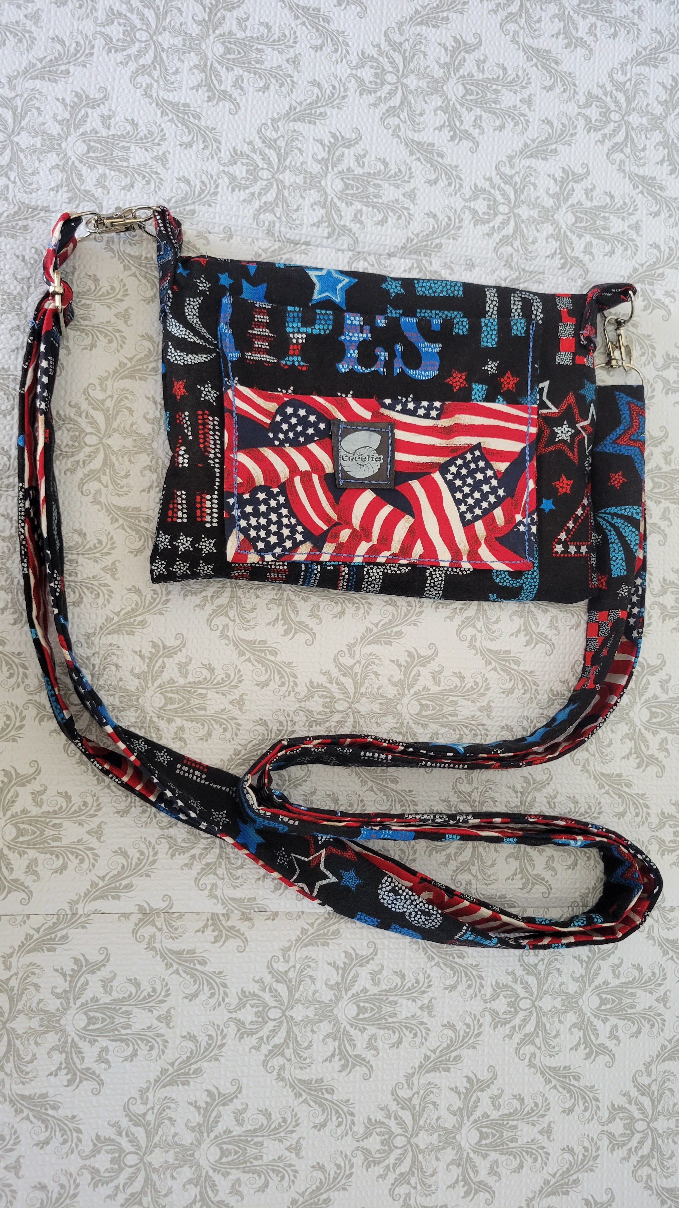 A patriotic-themed crossbody clutch purse with a black base and red, white, and blue American flag pattern, featuring a matching patterned shoulder strap, laid flat on a light beige damask-patterned fabric background.
