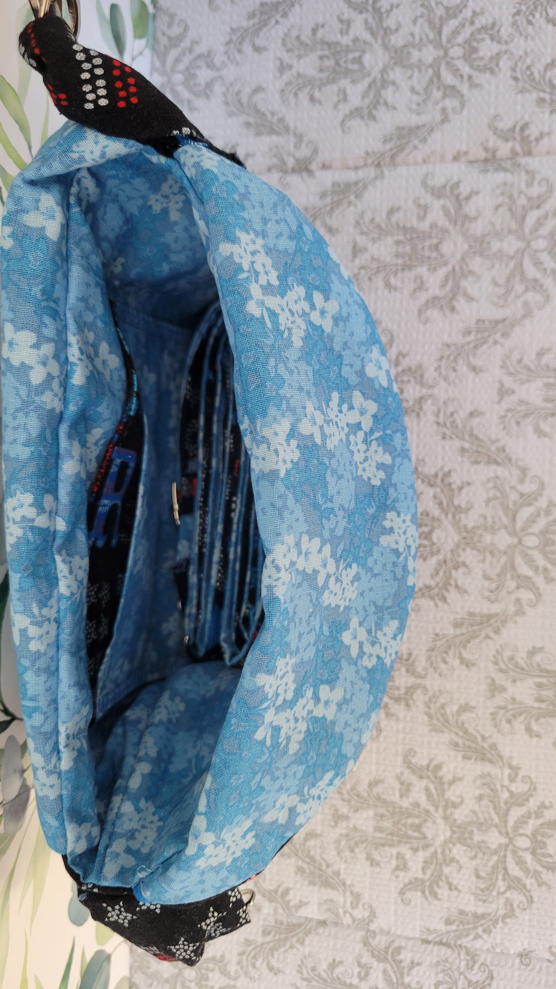 A blue purse with a white floral pattern, shown open to reveal its interior lining and a black patterned strap, set against a light gray damask-patterned background.