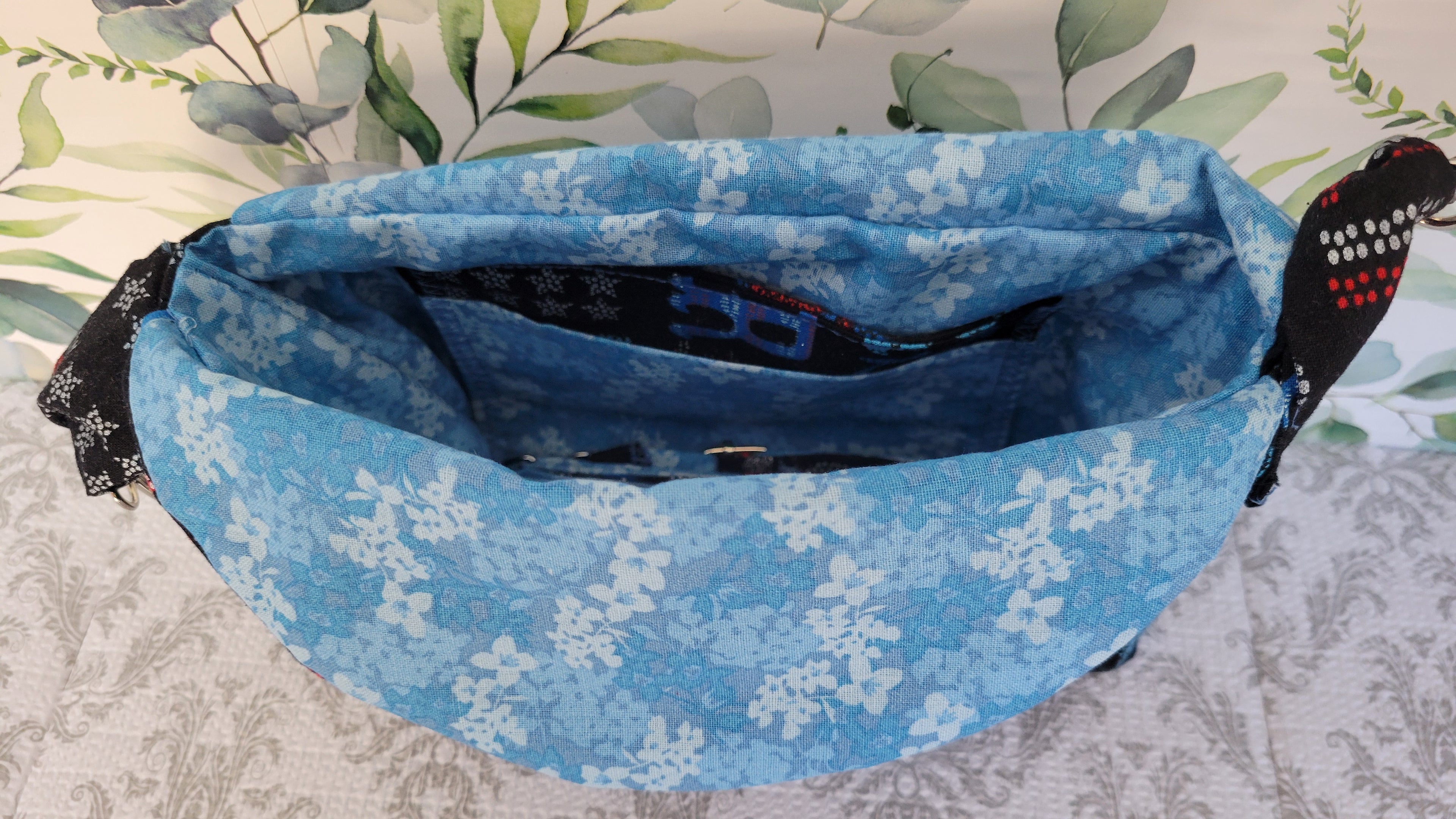 A blue waist purse with a white floral pattern, shown open to reveal its interior. The bag has black fabric side straps with white and red polka dots, set against a patterned light gray surface with a green leafy print background.