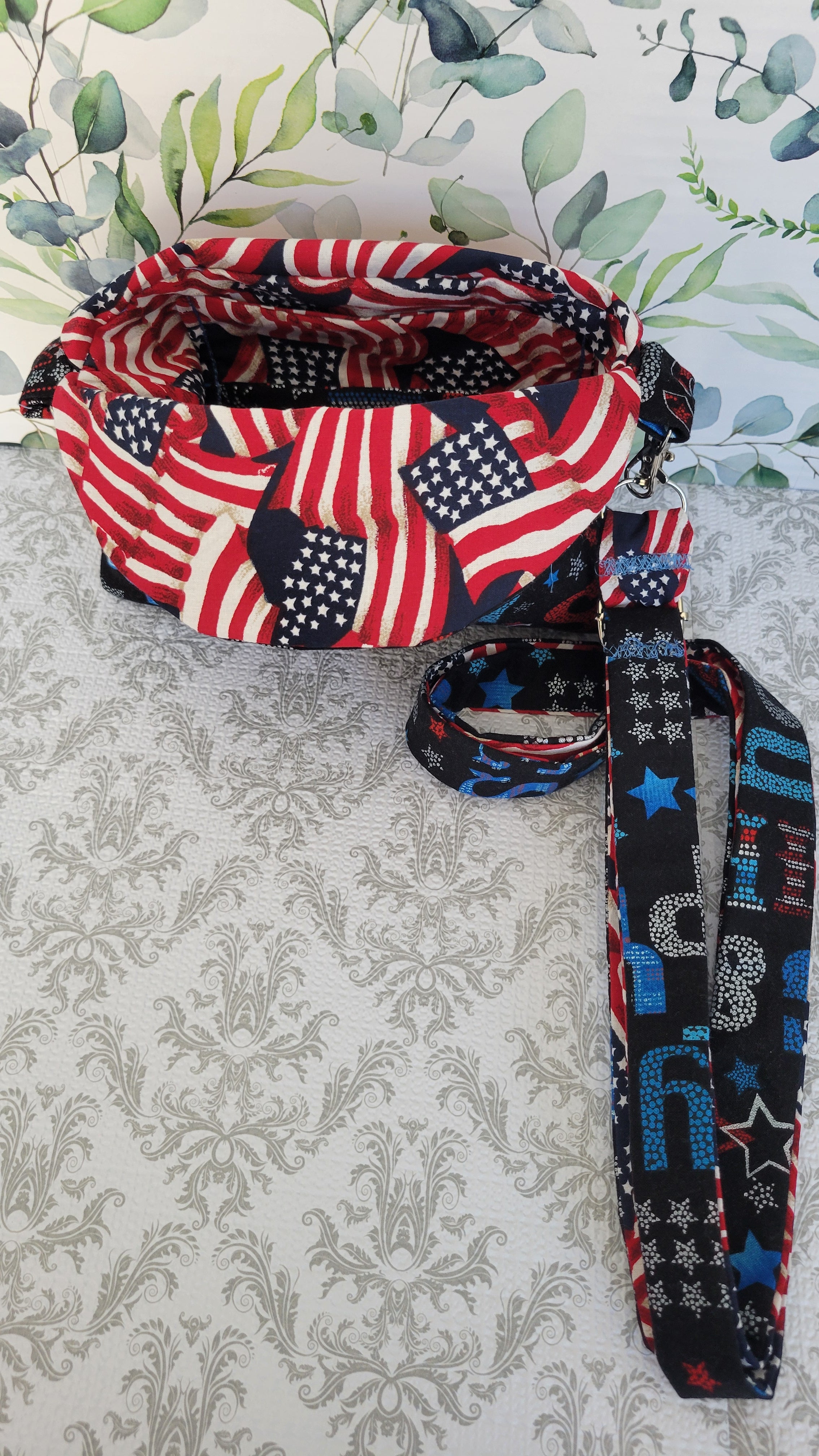 A patriotic-themed crossbody purse featuring a red, white, and blue American flag print on the exterior and a black wrist strap with blue and red text and star patterns, displayed on a gray damask fabric surface.