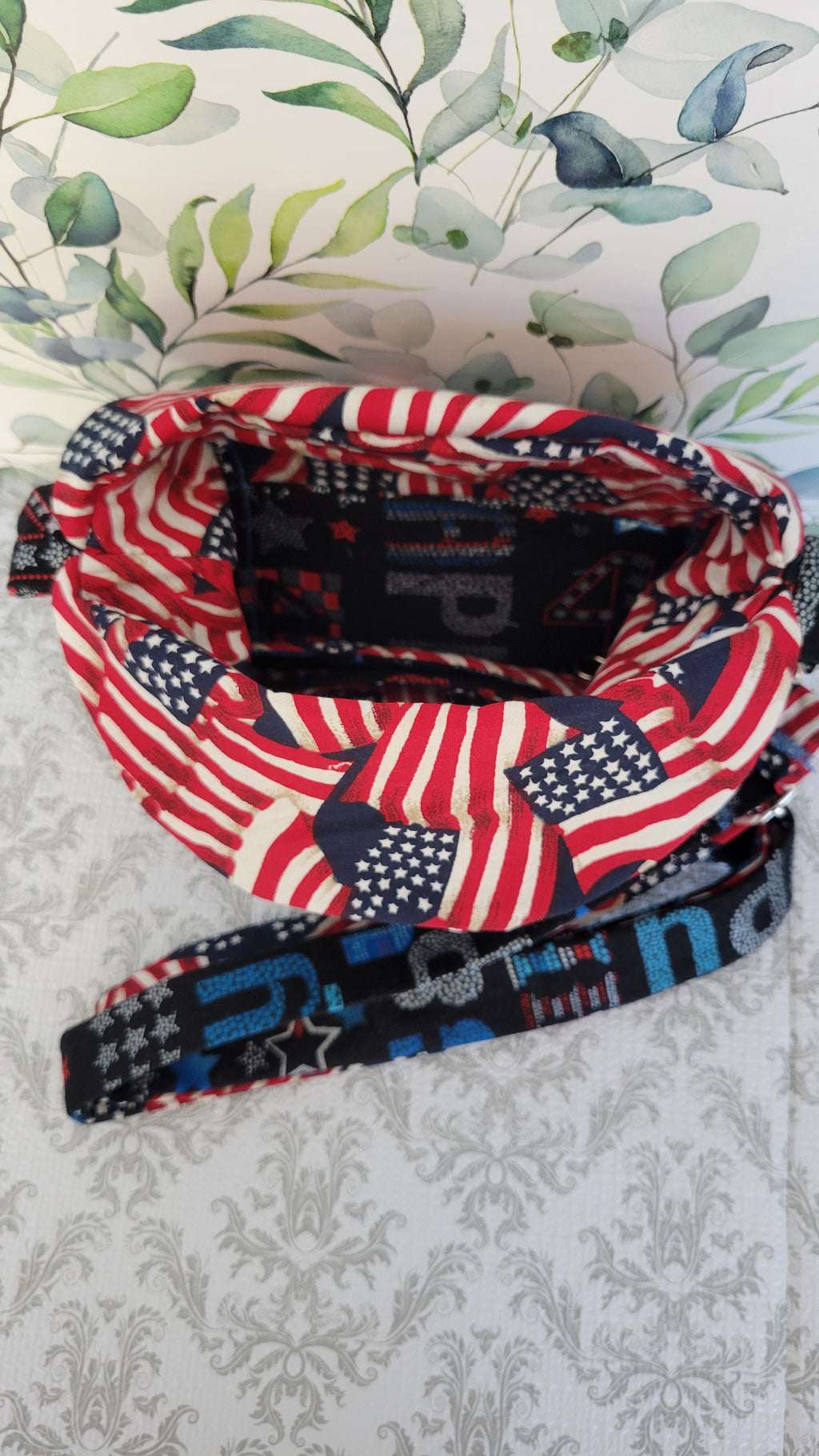 A purse with an American flag pattern on the front and a black inner band printed with 'Patriot' in blue, red, and white, displayed on a patterned surface.