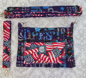 Multicolored fabric crossbody purse with American flag pattern on a textured gray background. Beside the purse is an adjustable strap, and a hand strap with the american flag pattern.