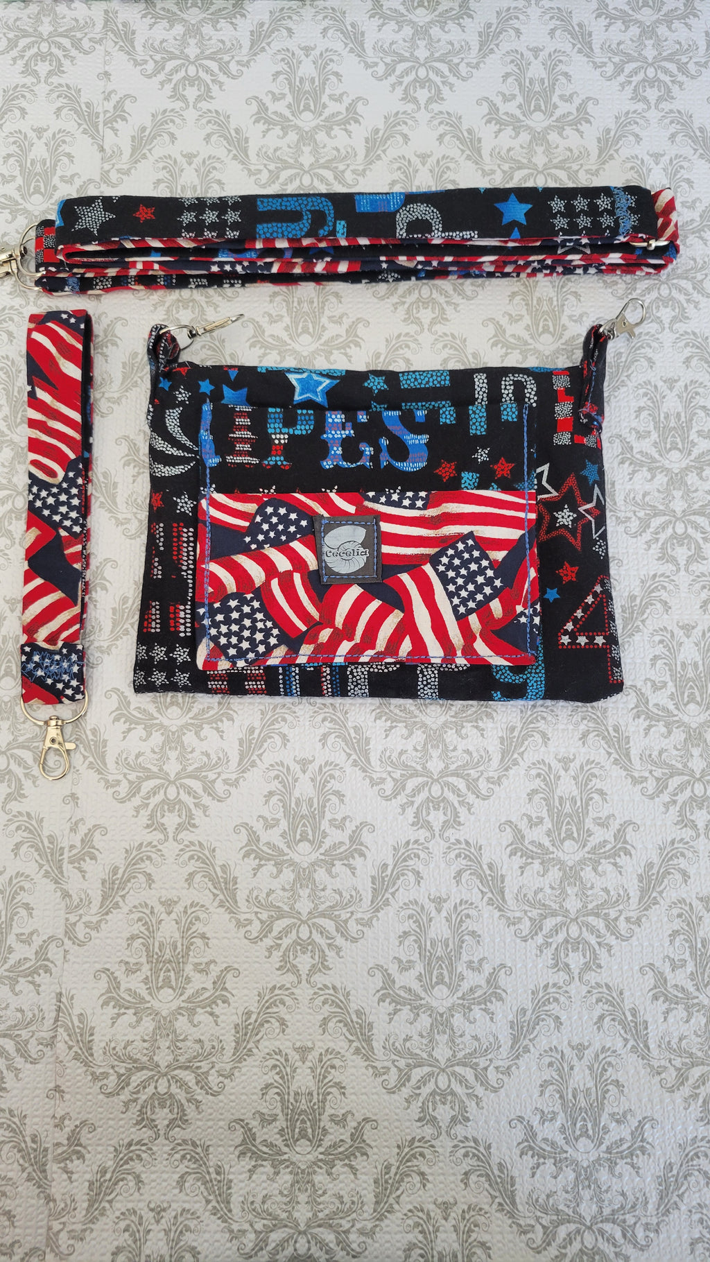A patriotic-themed fabric clutch purse with a matching wristlet strap, laid flat on a gray damask patterned background. The bag features a front pocket with a red, white, and blue American flag and stars-and-stripes print, and the strap has a black background with red and blue stars and stripes.