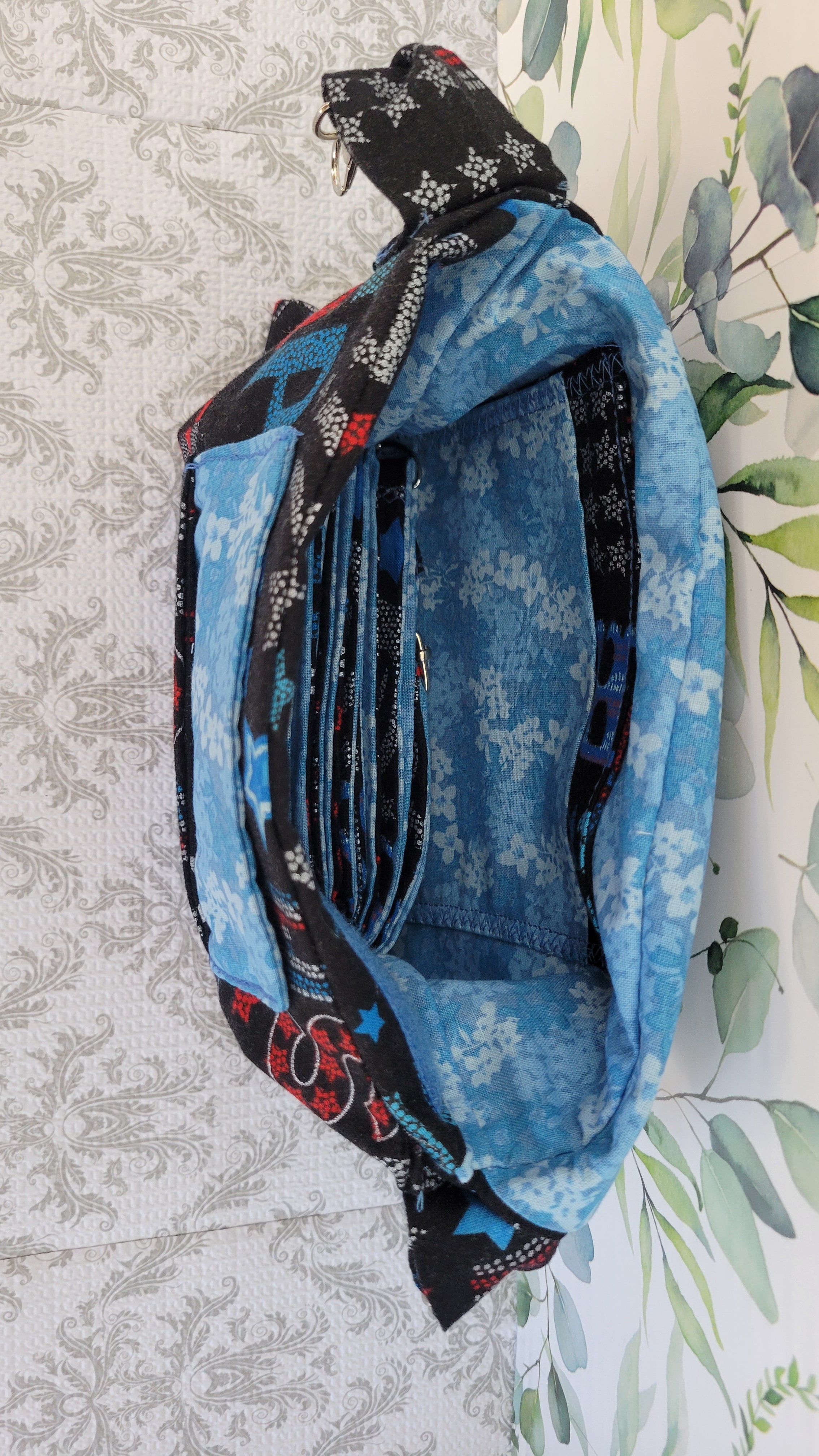 A handmade fabric crossbody bag with multiple pockets, featuring a blue floral print, a black and white patterned strap, and red accents, laid flat on a patterned background.