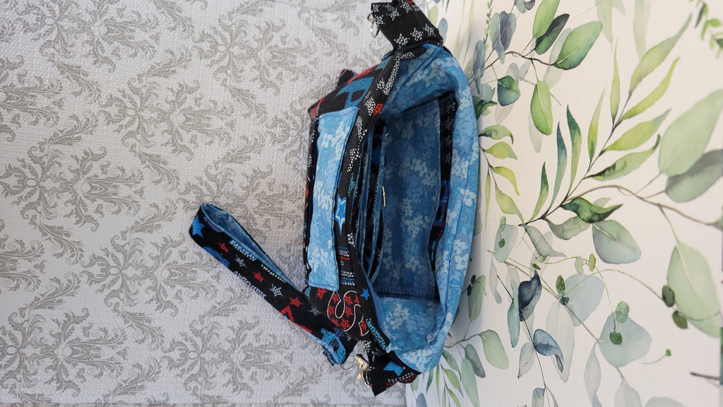 A blue patterned fabric bag with black and red patterned straps, laid flat on a white background with a gray damask pattern on the left and a green leaf print on the right. The purse is open to see two pockets in one inside the the purse.