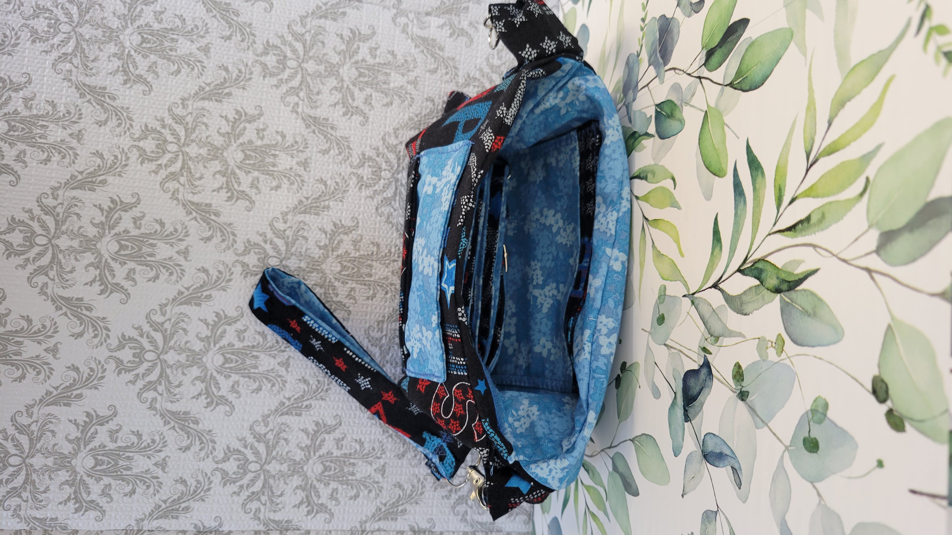 A blue patterned fabric bag with black and red patterned straps, laid flat on a white background with a gray damask pattern on the left and a green leaf print on the right. The purse is open to see two pockets in one inside the the purse.