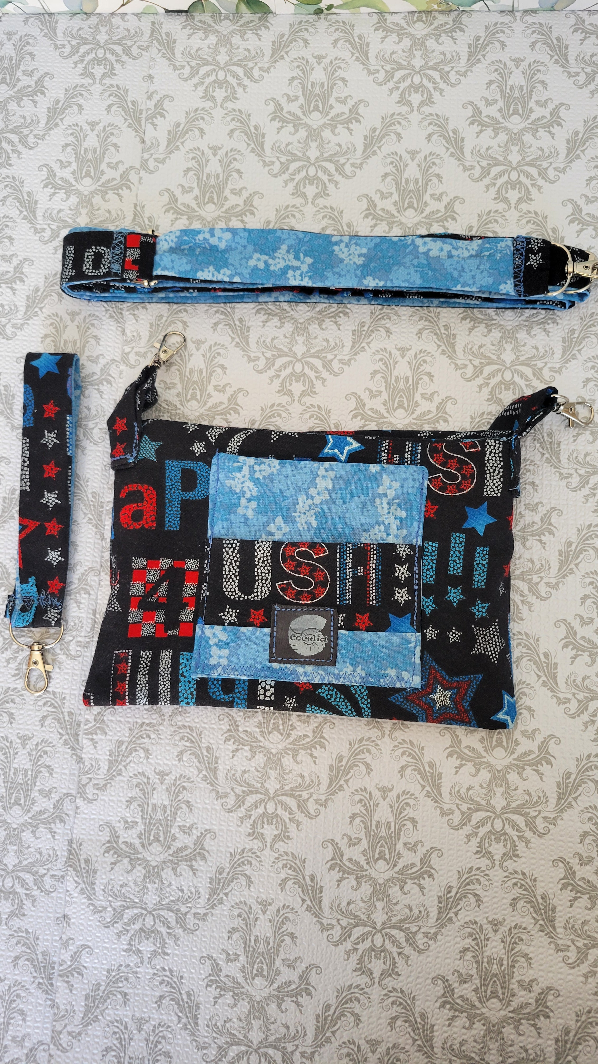 A handmade patchwork fabric purse and matching wristlet strap laid flat on a beige damask-patterned surface. The pouch and strap feature a patchwork design of light blue, black, red, and white fabrics with letters and stars.
