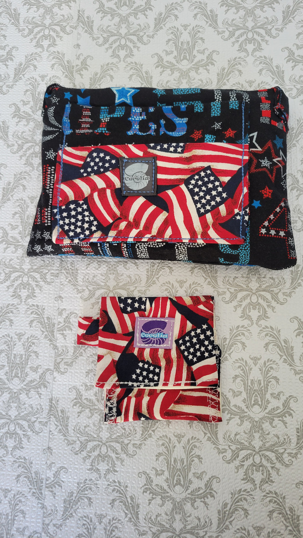 A wallet with a patriotic American flag print, shown both unfolded and folded on a light gray damask-patterned fabric background. The purse features red, white, and blue flag designs with a central black patch containing a white oval logo.