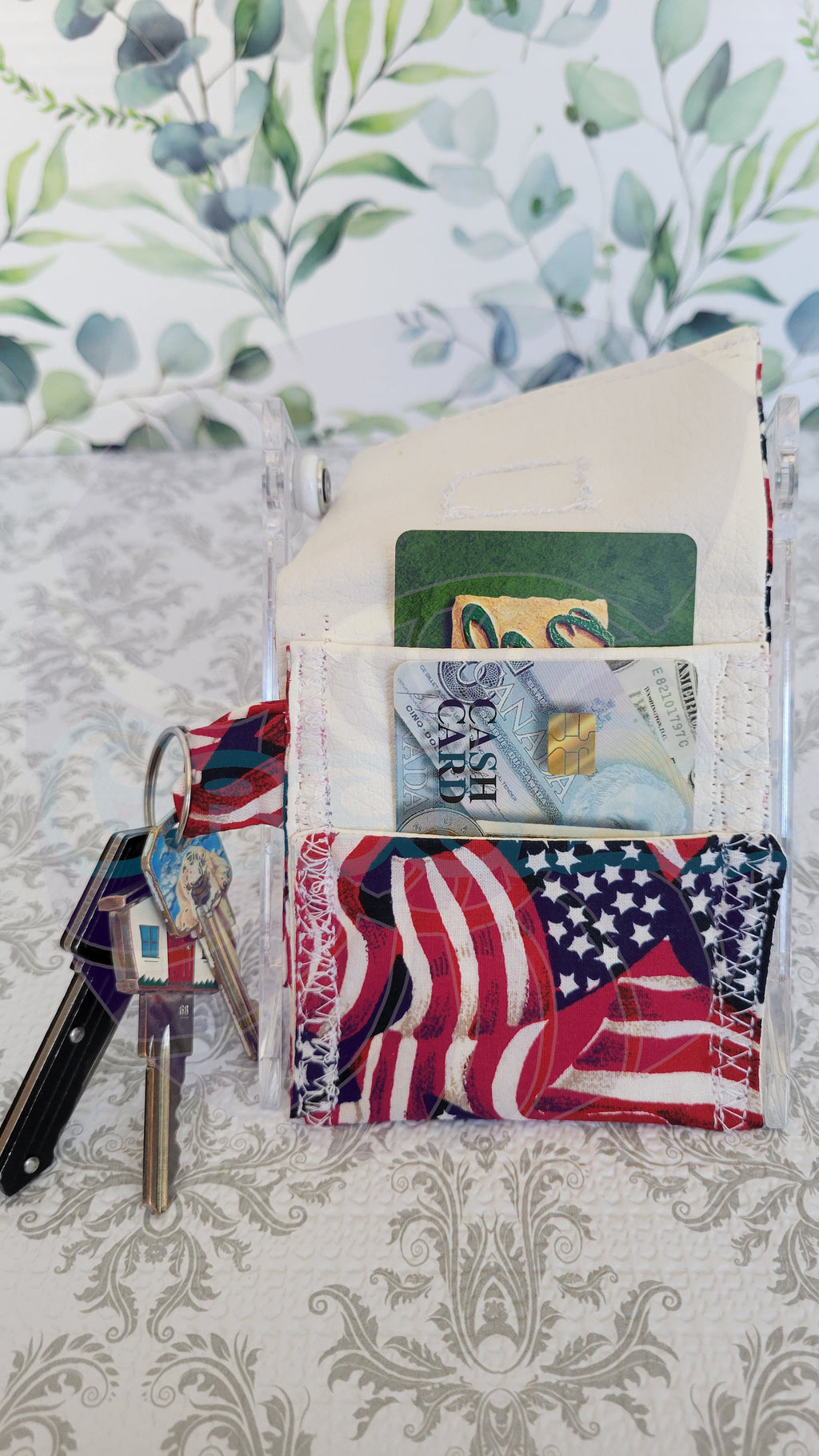 A clear acrylic wallet with a patriotic American flag patterned fabric lining, shown holding cash, cards, and keys on a patterned tablecloth.