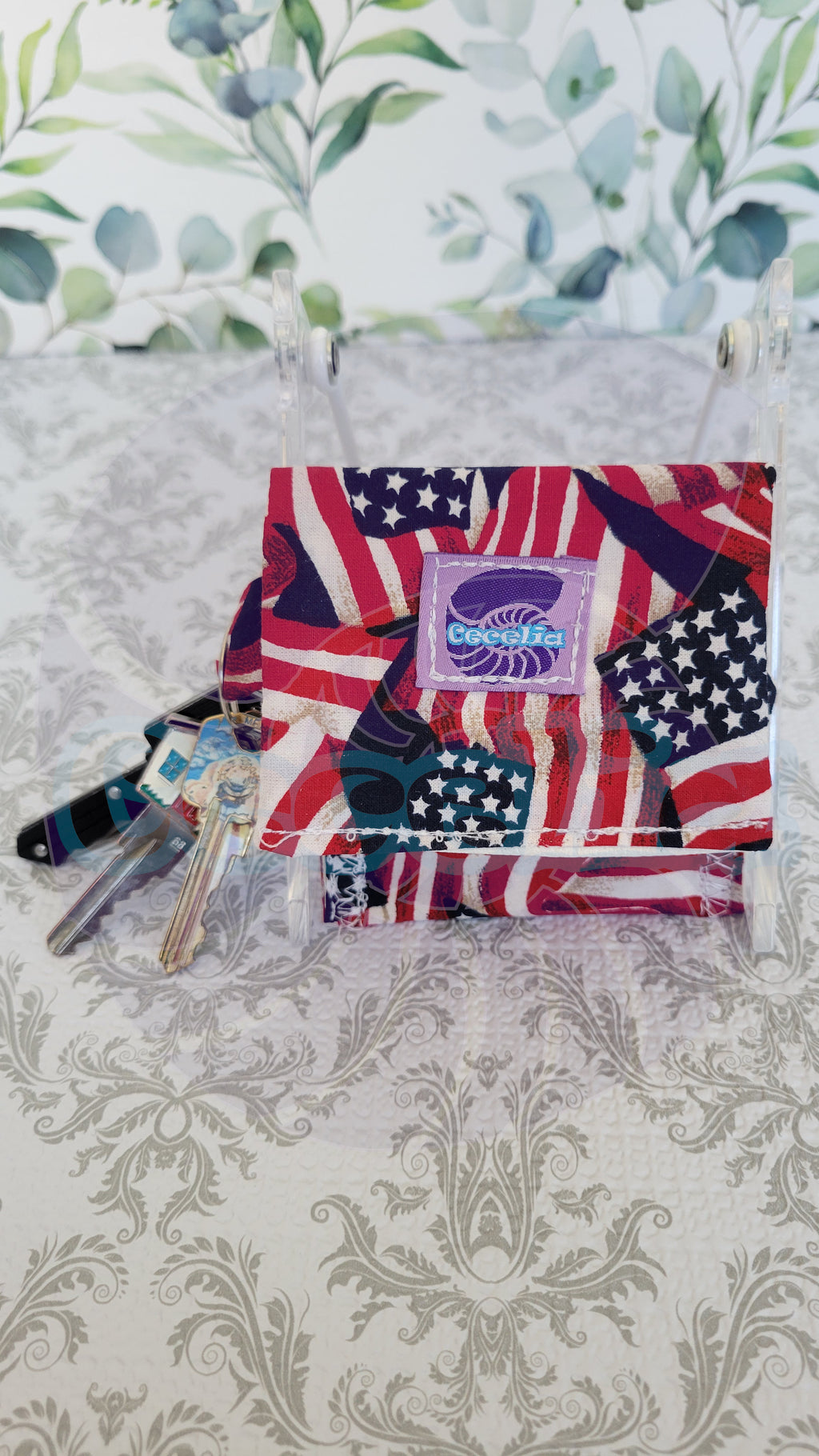 A wallet with an American flag patchwork print and a central purple patch reading 'Patriotic', displayed on a clear acrylic stand on a patterned tablecloth with floral wallpaper in the background