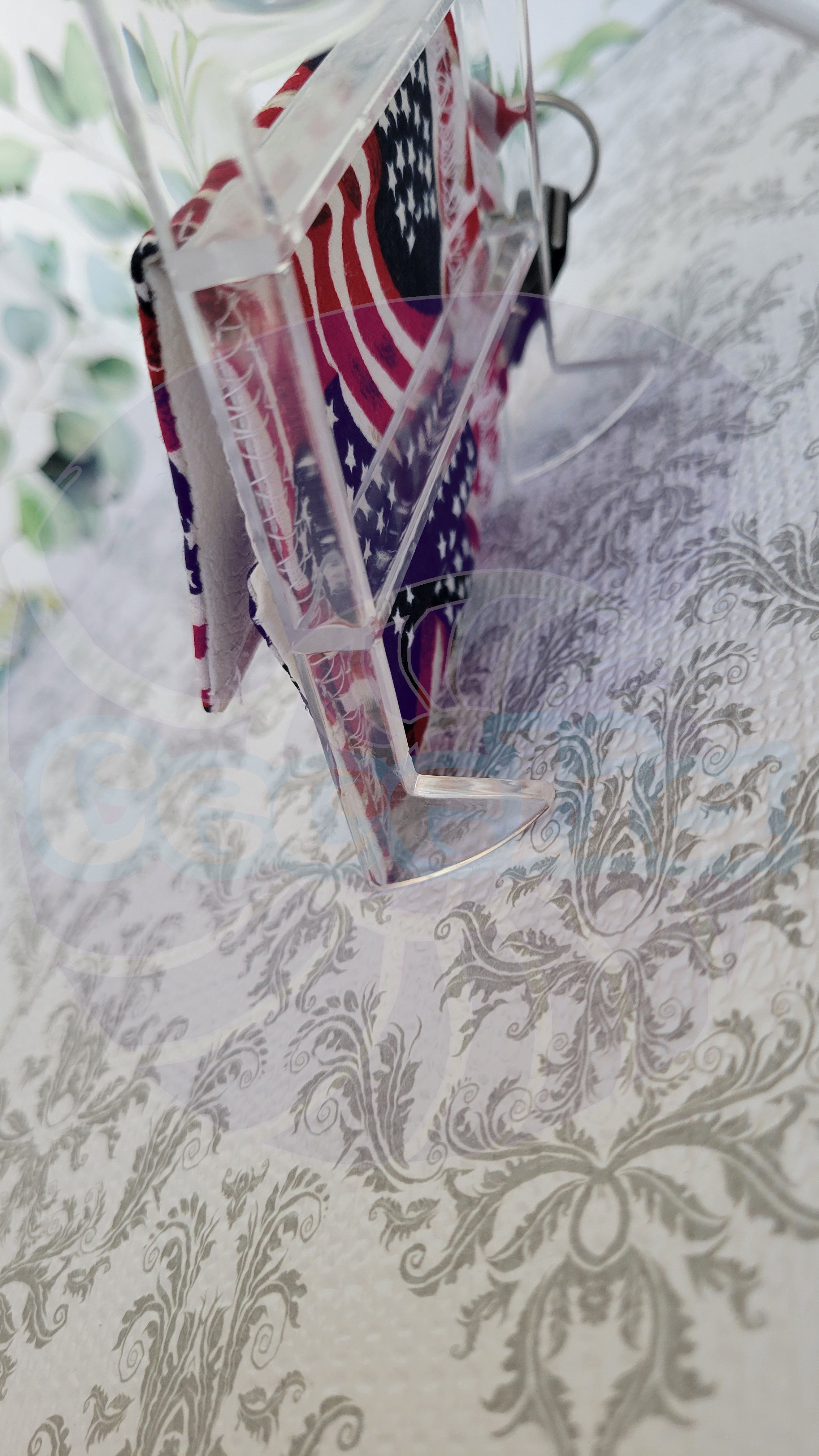 A clear acrylic stand holding a small printed wallet with an American flag pattern, photographed on a light patterned surface.