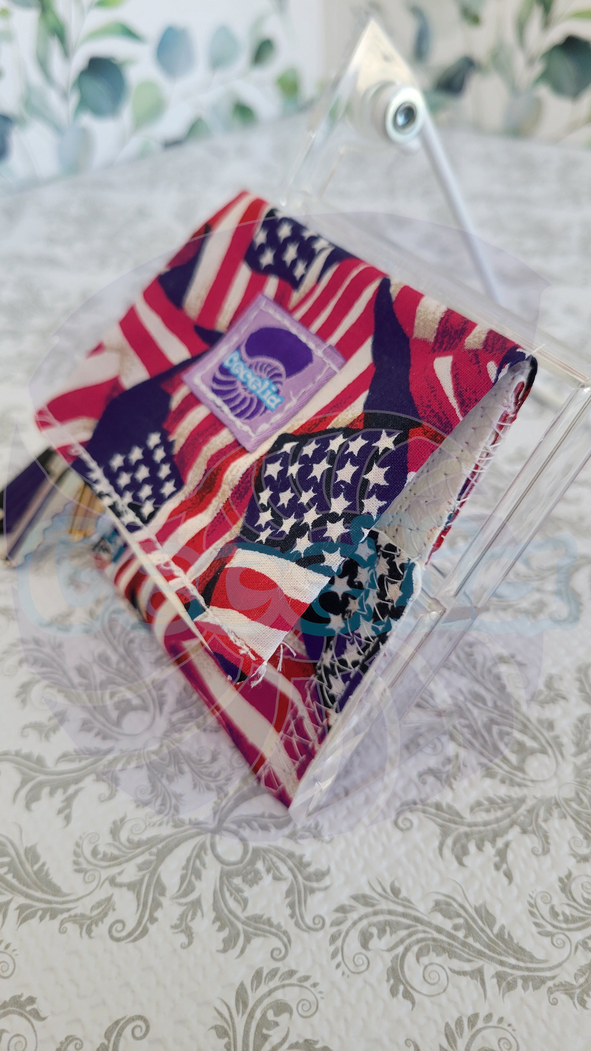 A clear acrylic stand holding a wallet  item with an American flag pattern in red, white, and blue, photographed on a patterned light surface.
