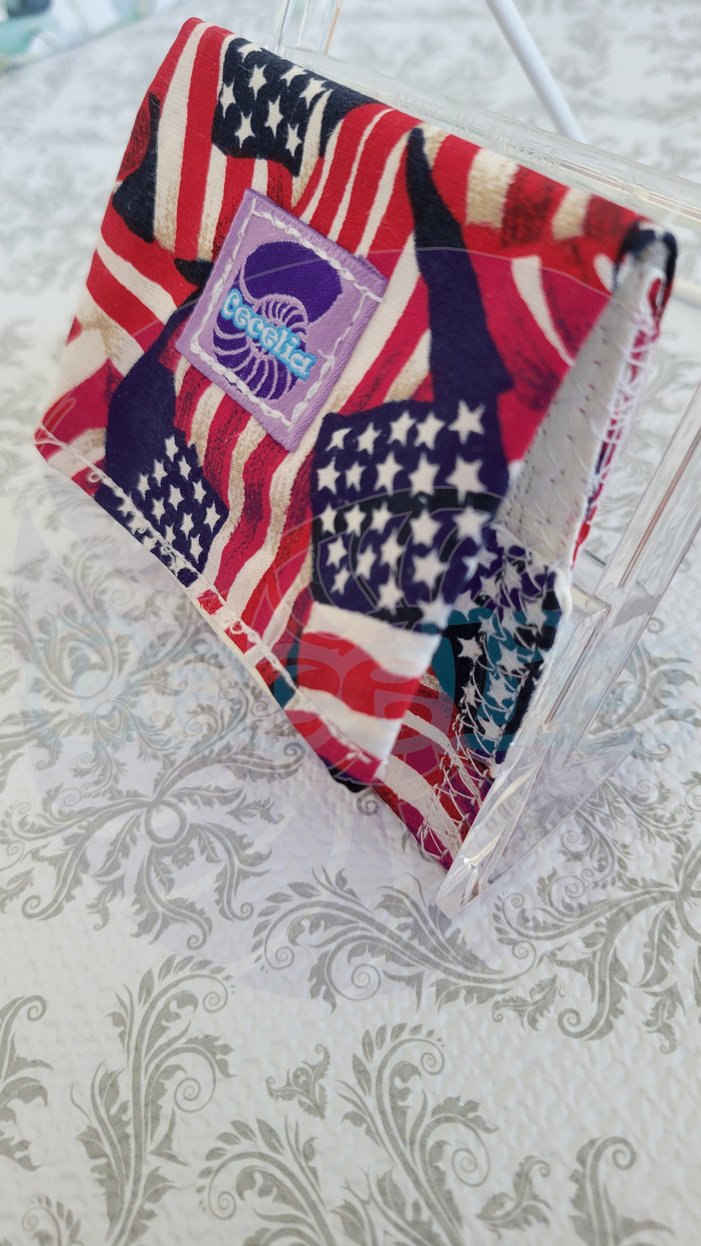 A wallet with a patriotic American flag print featuring red, white, and blue stripes and stars, folded inside a clear plastic bag. A purple square patch on the mask displays the brand name 'The Jellyfish'.