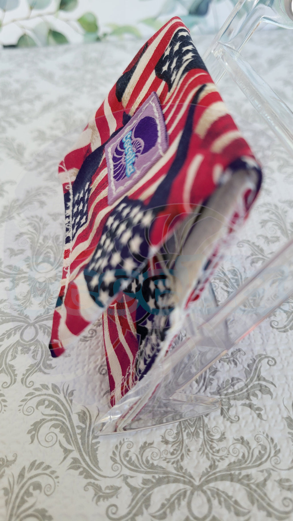 A wallet with an American flag-inspired design in red, white, and blue, displayed on a clear stand over a damask-patterned surface.
