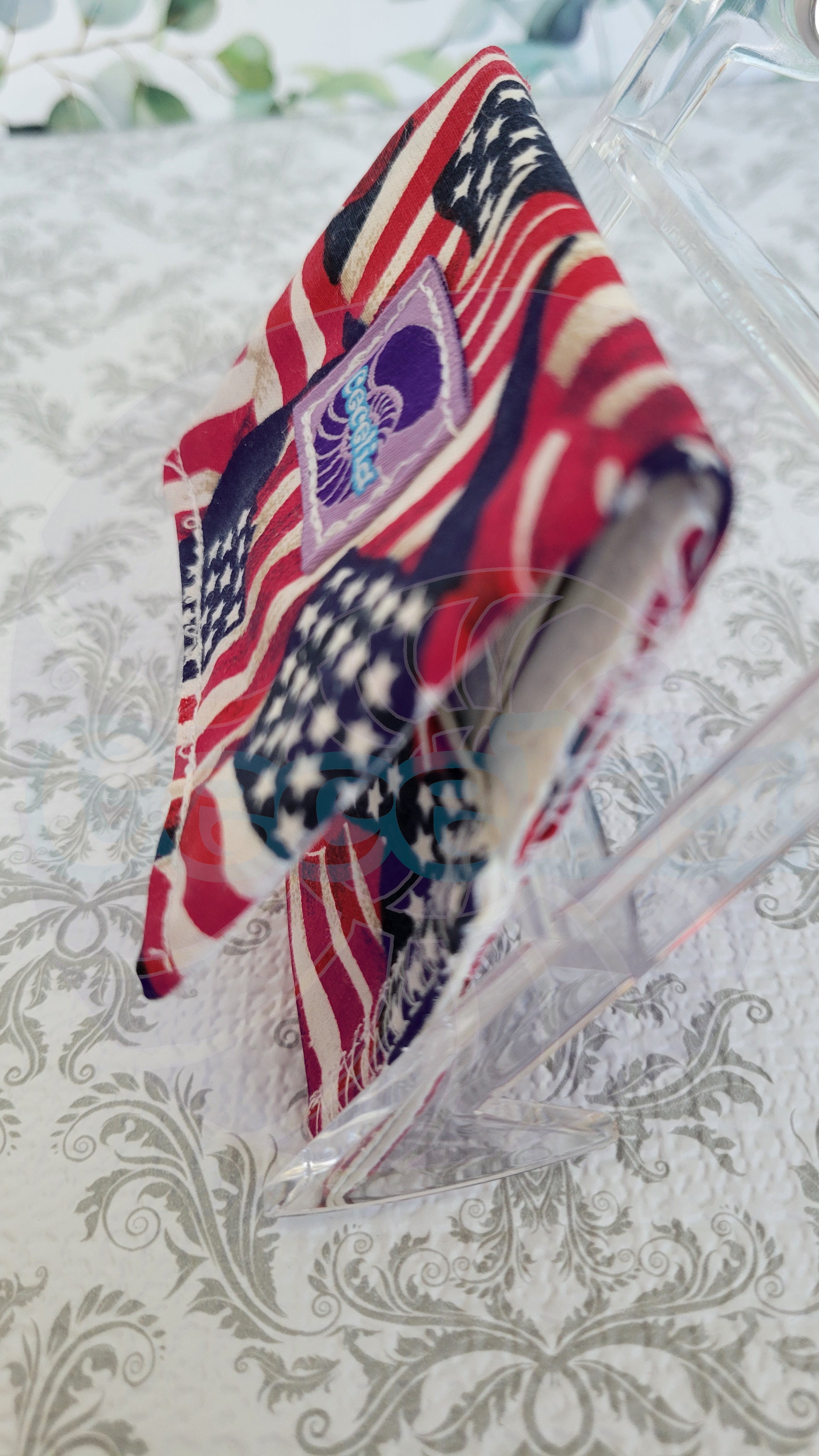 A wallet with an American flag-inspired design in red, white, and blue, displayed on a clear stand over a damask-patterned surface.