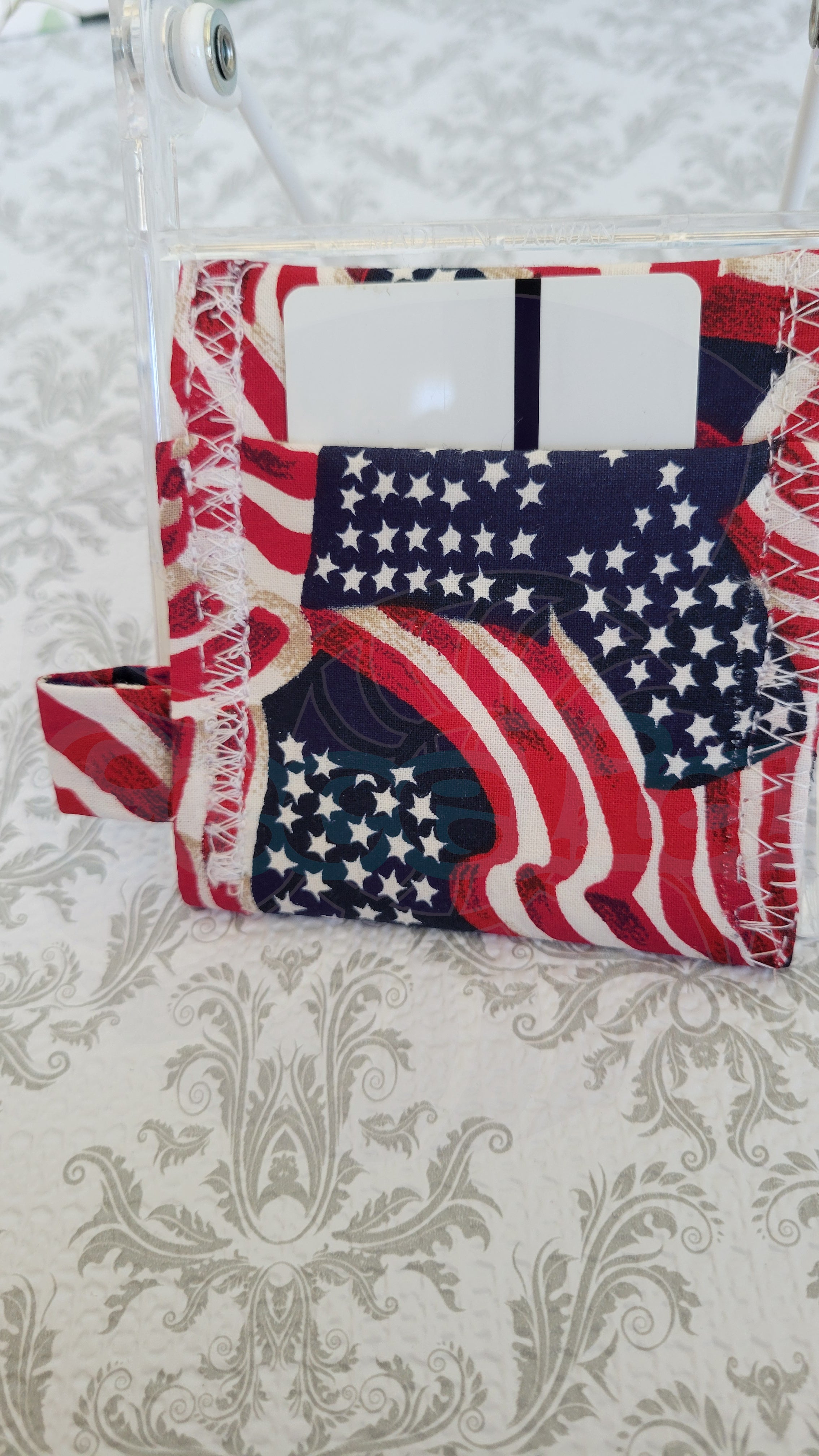 A patriotic-themed wallet featuring a red, white, and blue American flag pattern, shown with a white card inserted into a clear plastic sleeve on a patterned beige and gray damask surface.
