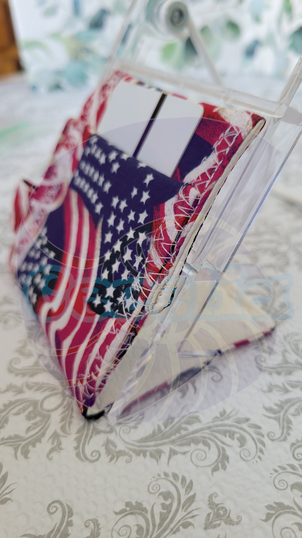 A small wallet accessory with an American flag print, shown inside a clear plastic display stand on a patterned white surface.