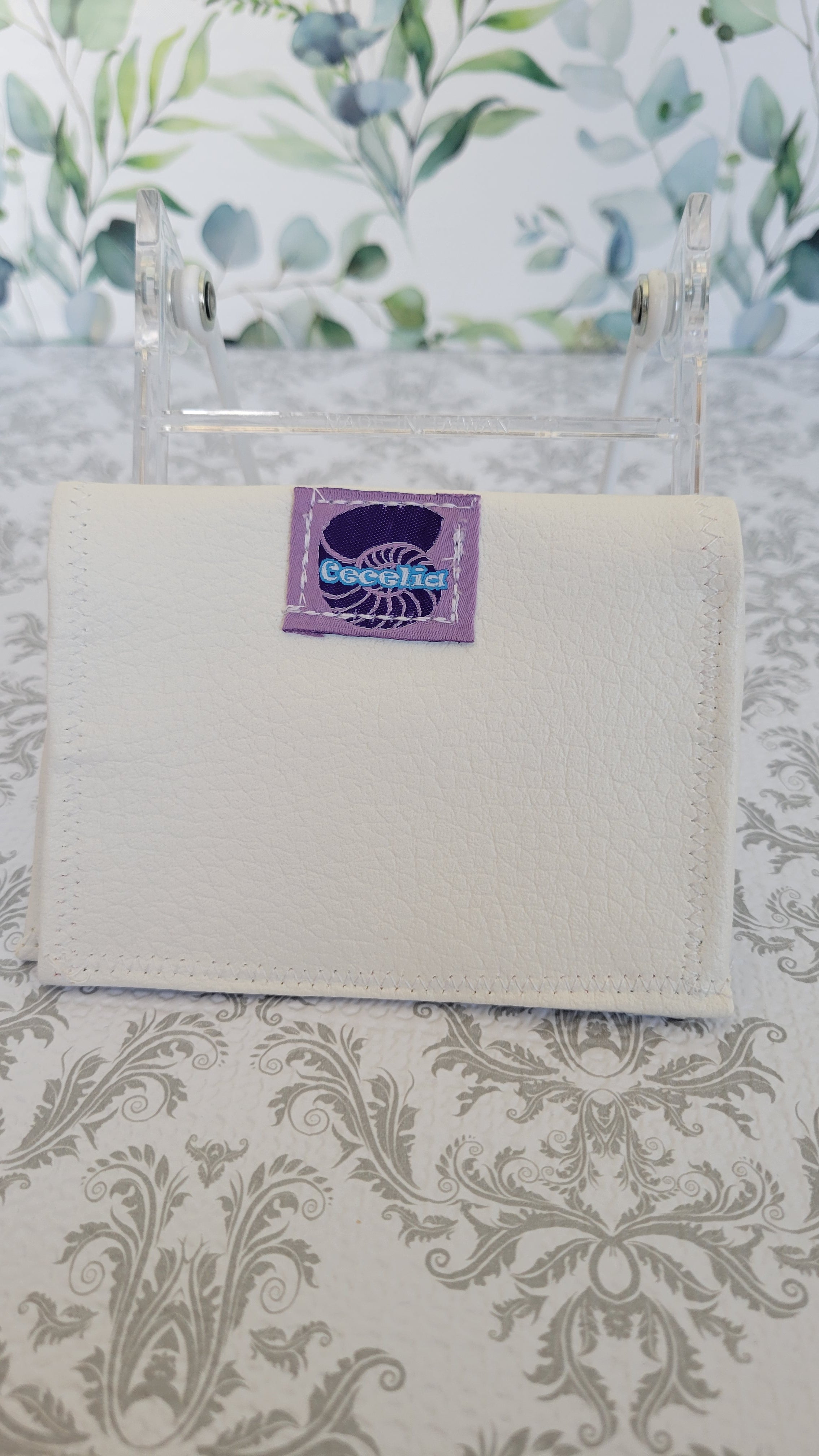 An off-white textured leather wallet with a purple brand patch reading 'Cacelia', displayed on a clear acrylic stand over a patterned beige and gray damask surface.