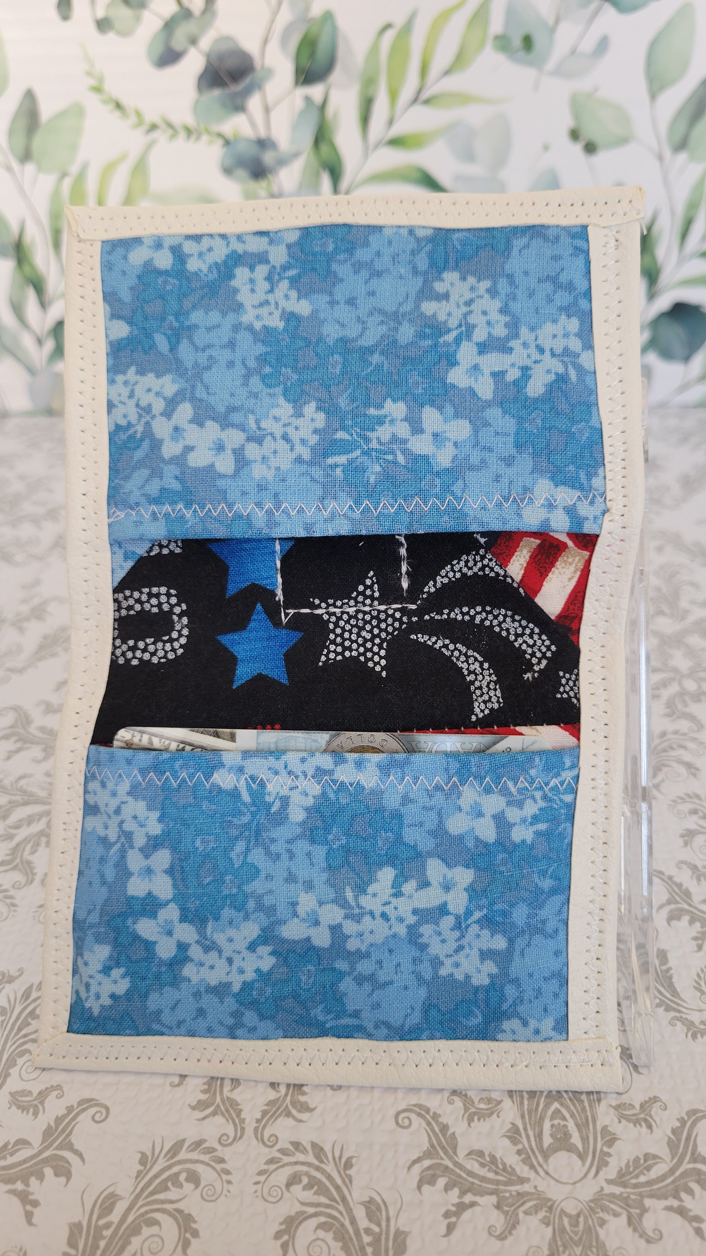 A handmade fabric wallet with a patriotic theme, featuring blue floral patterns, a black section with white stars, and a small red, white, and blue flag accent. The pouch is displayed on a patterned beige surface.