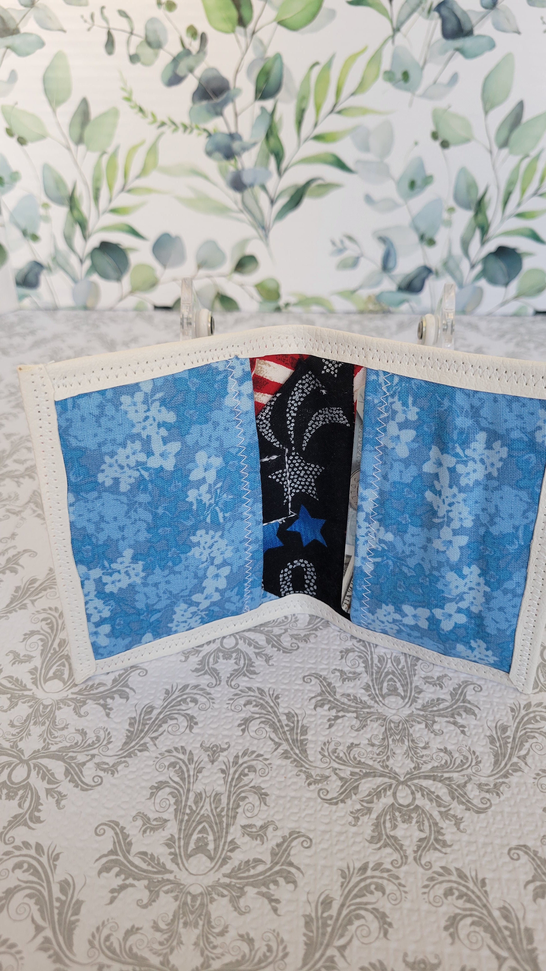 A handmade wallet with a patriotic theme, featuring two blue floral panels and a central black panel with stars and stripes, displayed on a patterned surface.