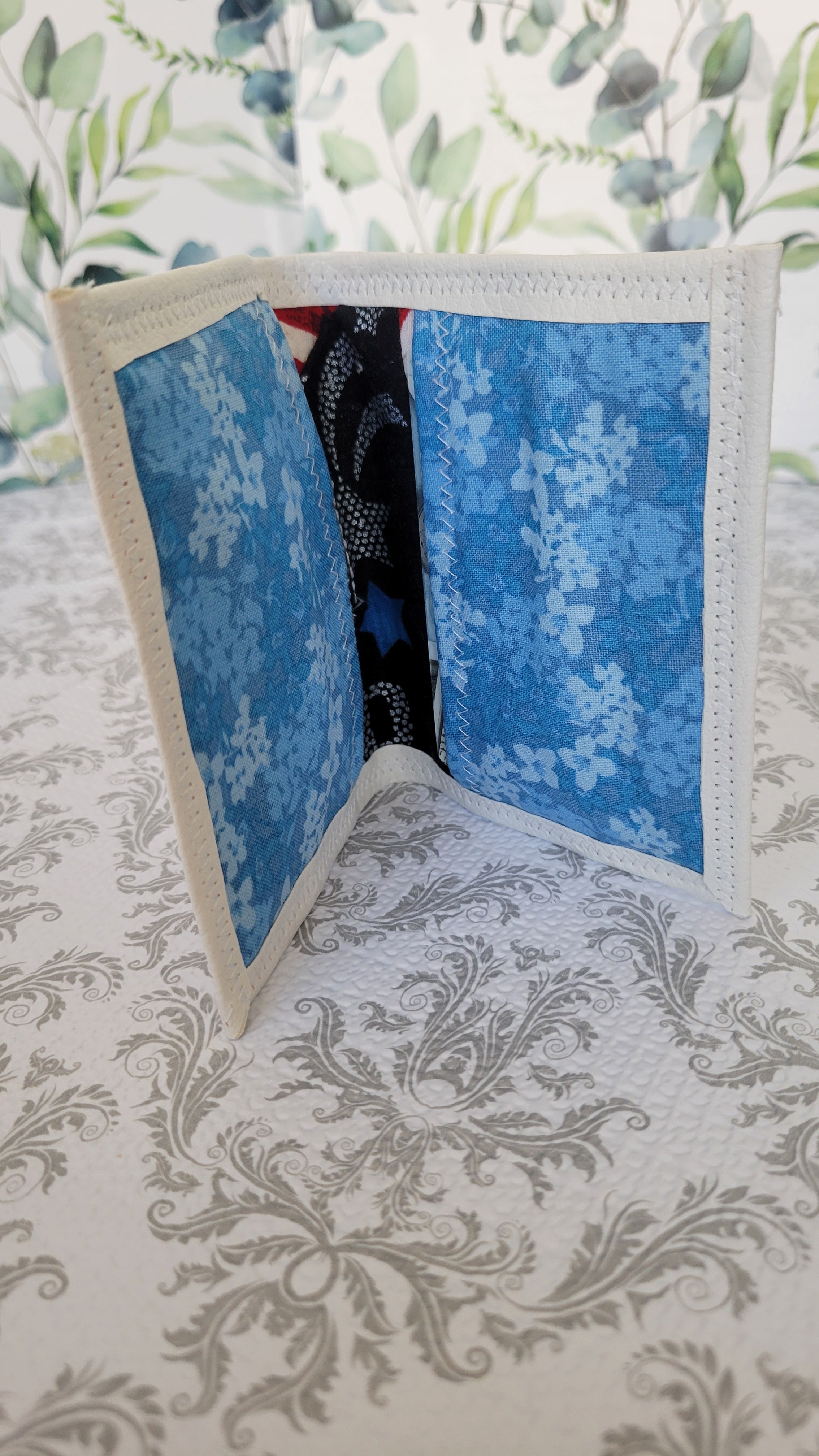 A handmade bifold wallet with a cream-colored fabric trim and blue patterned fabric lining, displayed open on a gray damask-patterned surface.