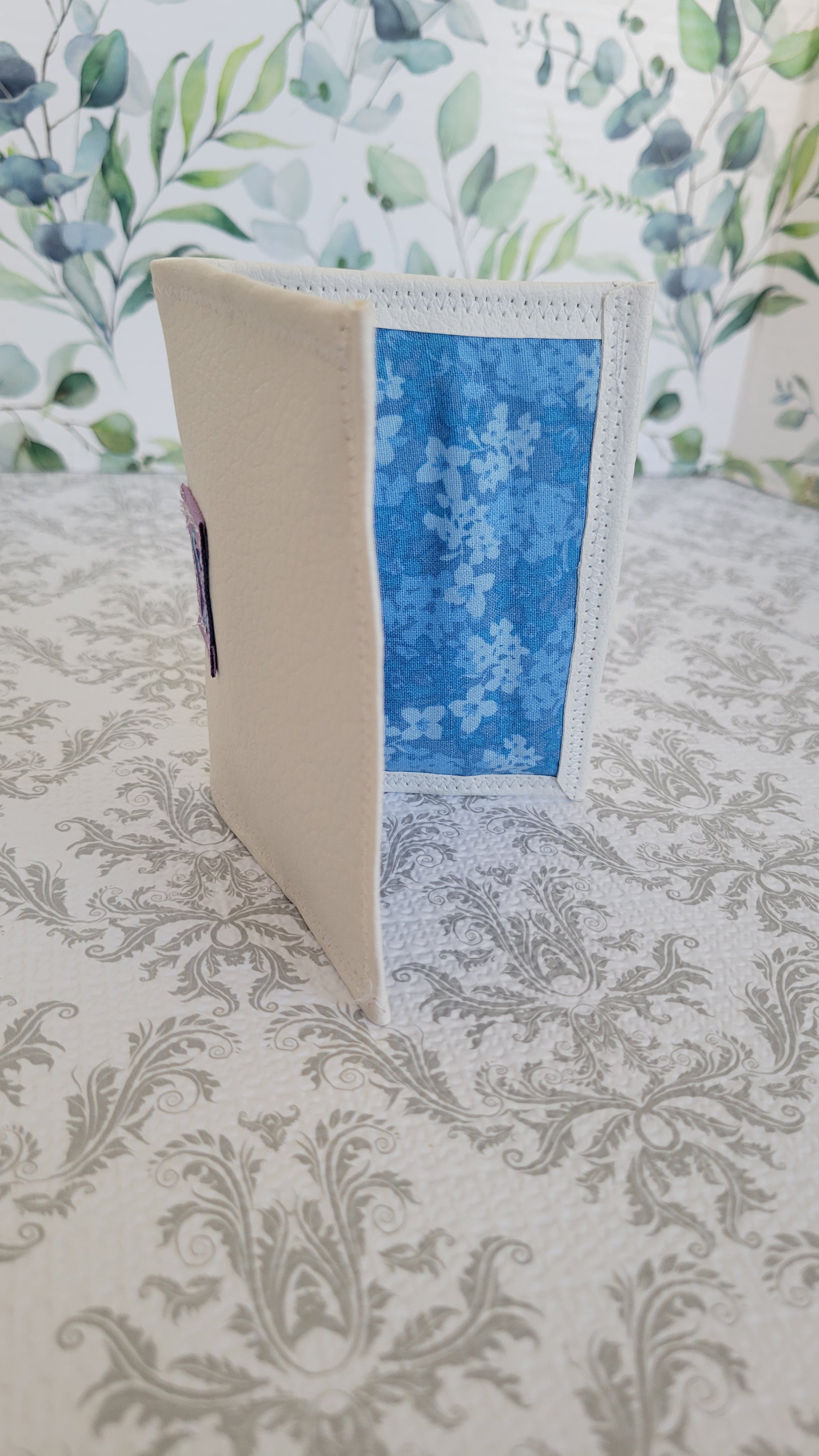 A handmade felt wallet standing upright on a patterned tablecloth, featuring a cream-colored exterior and a blue floral patterned interior pocket.