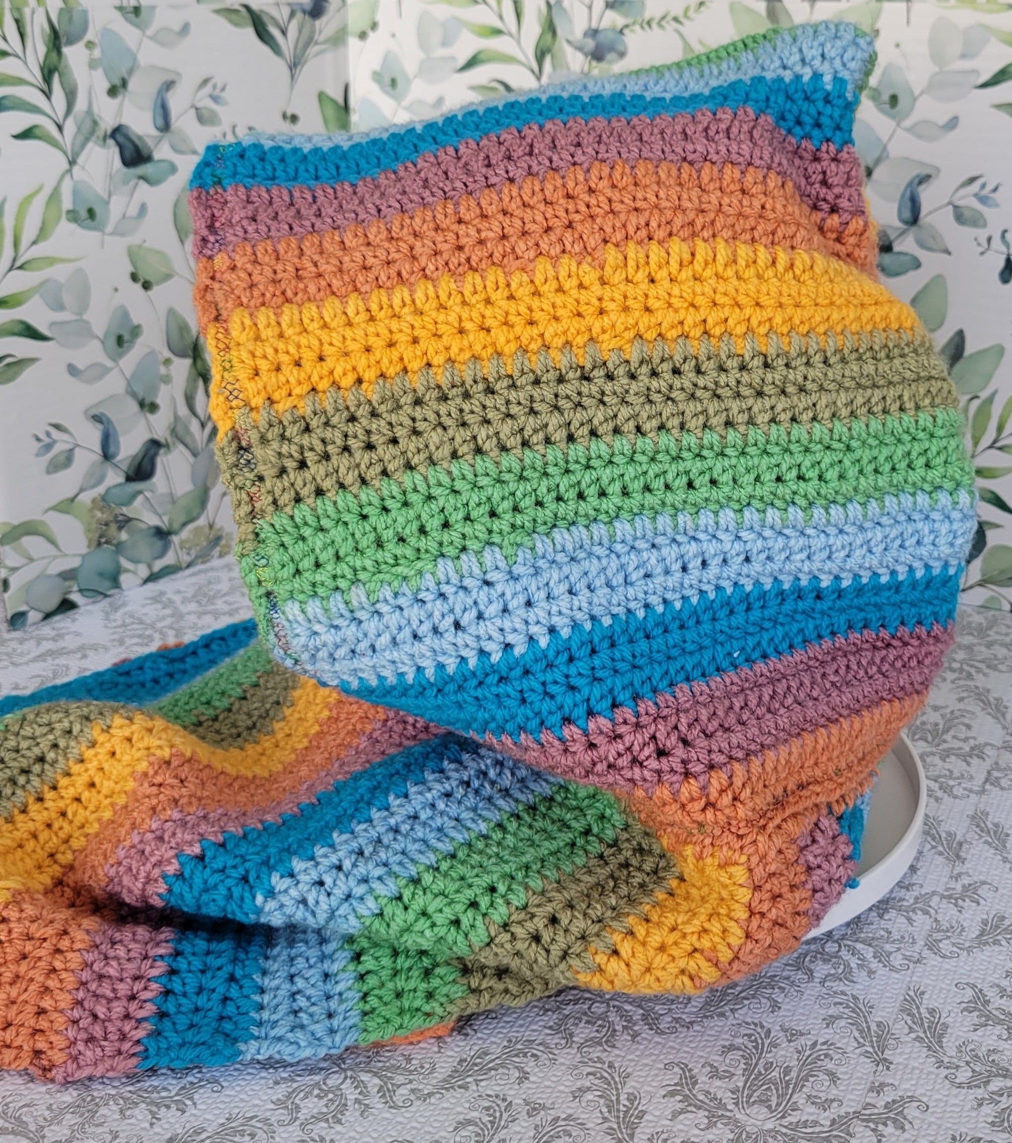 Colorful strips of crocheted scarf and hoodie with a white mannequin inside on a sky blue surface. The mannequin is facing the left side.