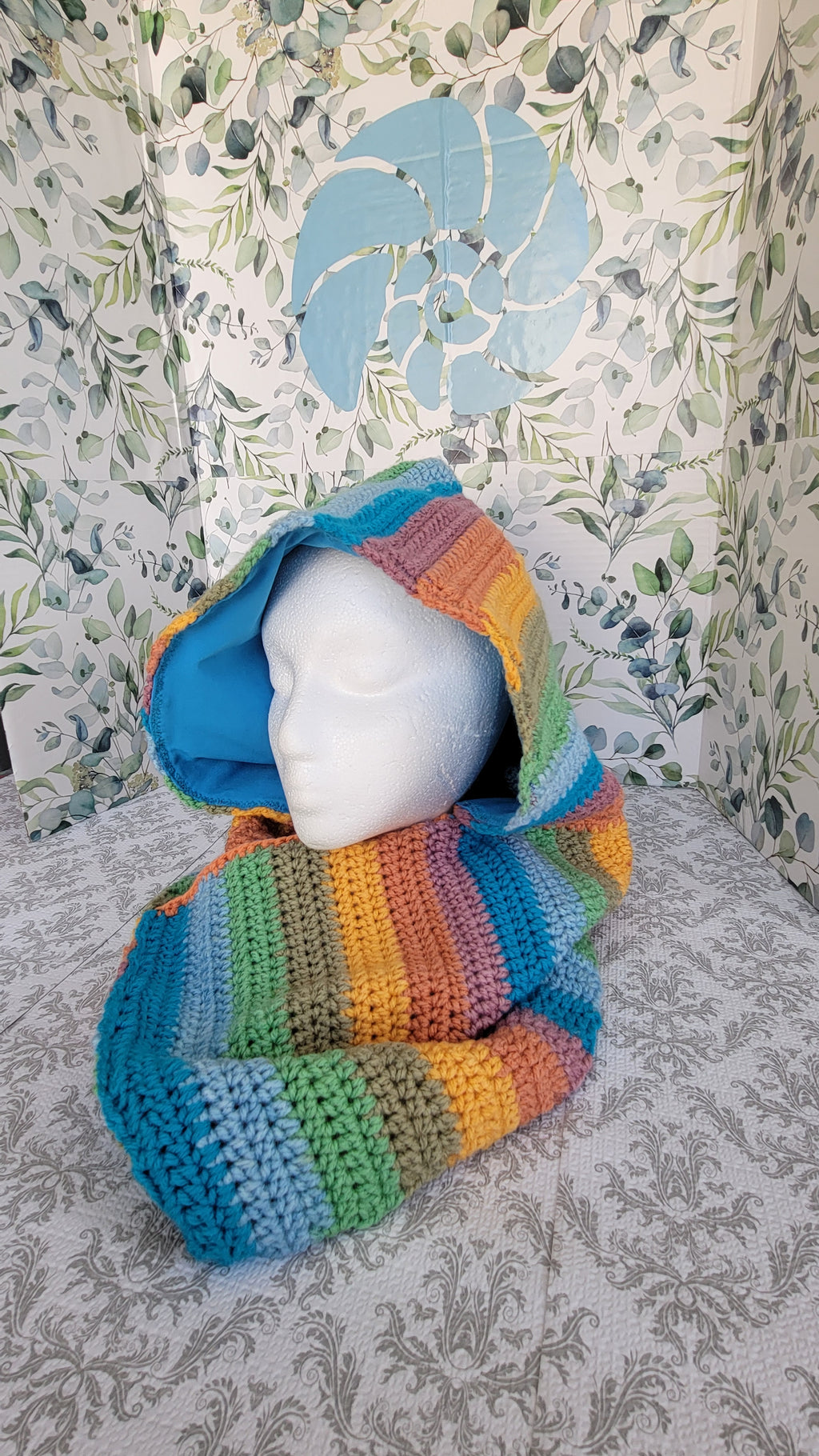 Colorful strips of crocheted scarf and hoodie with a white mannequin inside on a sky blue surface. The mannequin is facing forward.