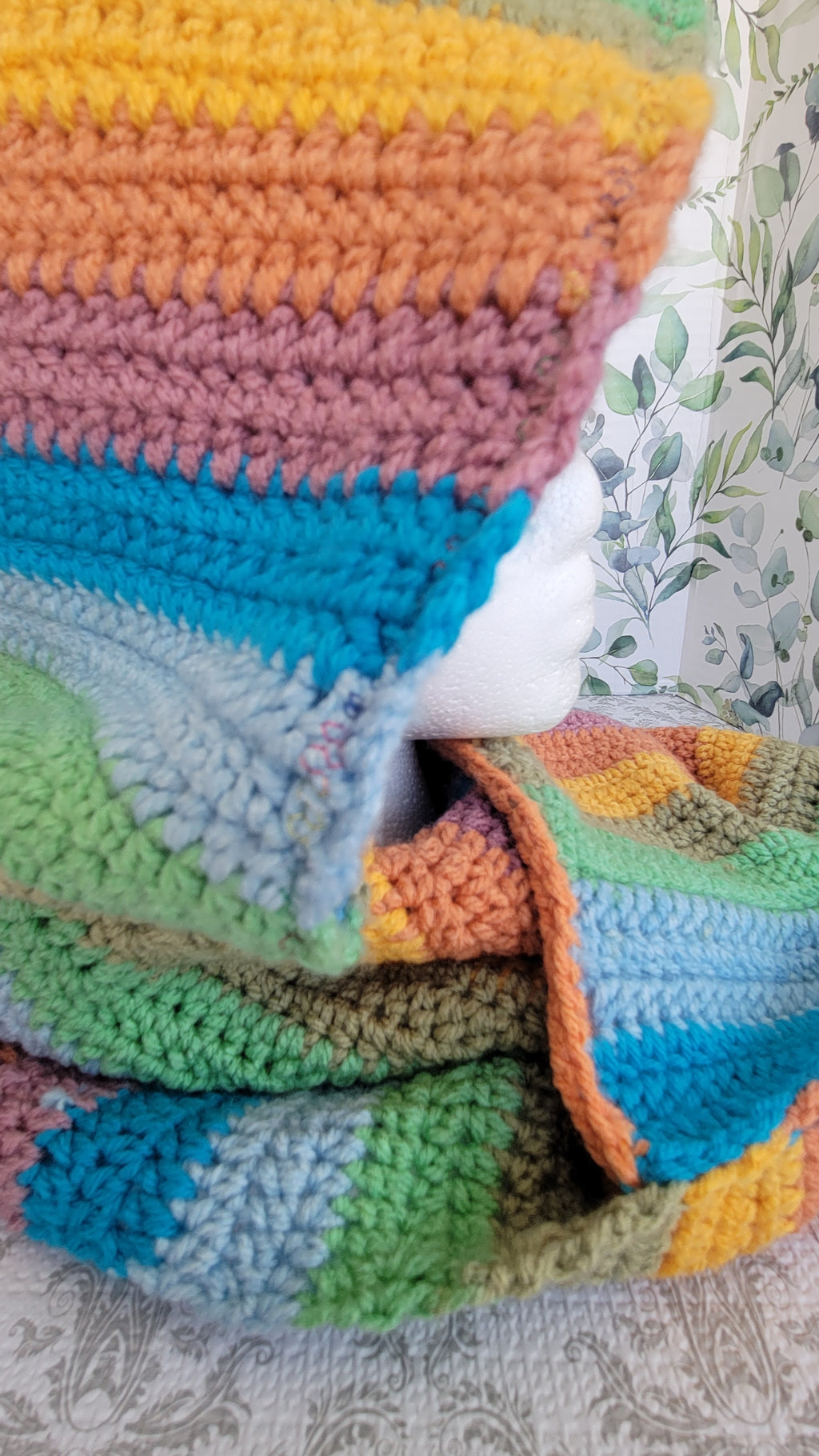 Colorful strips of crocheted scarf and hoodie with a white mannequin inside on a sky blue surface. The mannequin is facing left side.